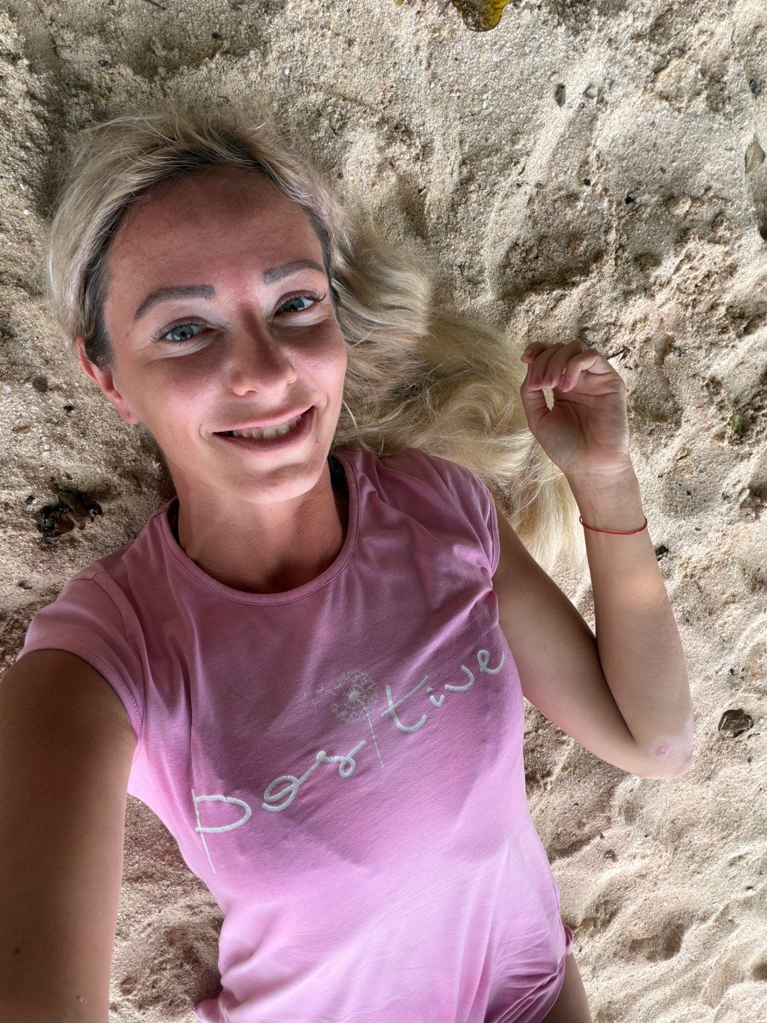 A woman with blonde hair is lying on a sandy beach, looking up at the camera and smiling. She is wearing a pink t-shirt with the word "Positive" written on it in white letters, and a small dandelion graphic above the word. A thin red bracelet is visible on her right wrist. The sand is light brown and textured, with a few small pebbles and dark debris scattered throughout. The background is out of focus, but appears to be more sand and possibly some foliage. This image was likely taken on a beach in Malay, Philippines, known for its popular tourist destinations like Boracay.