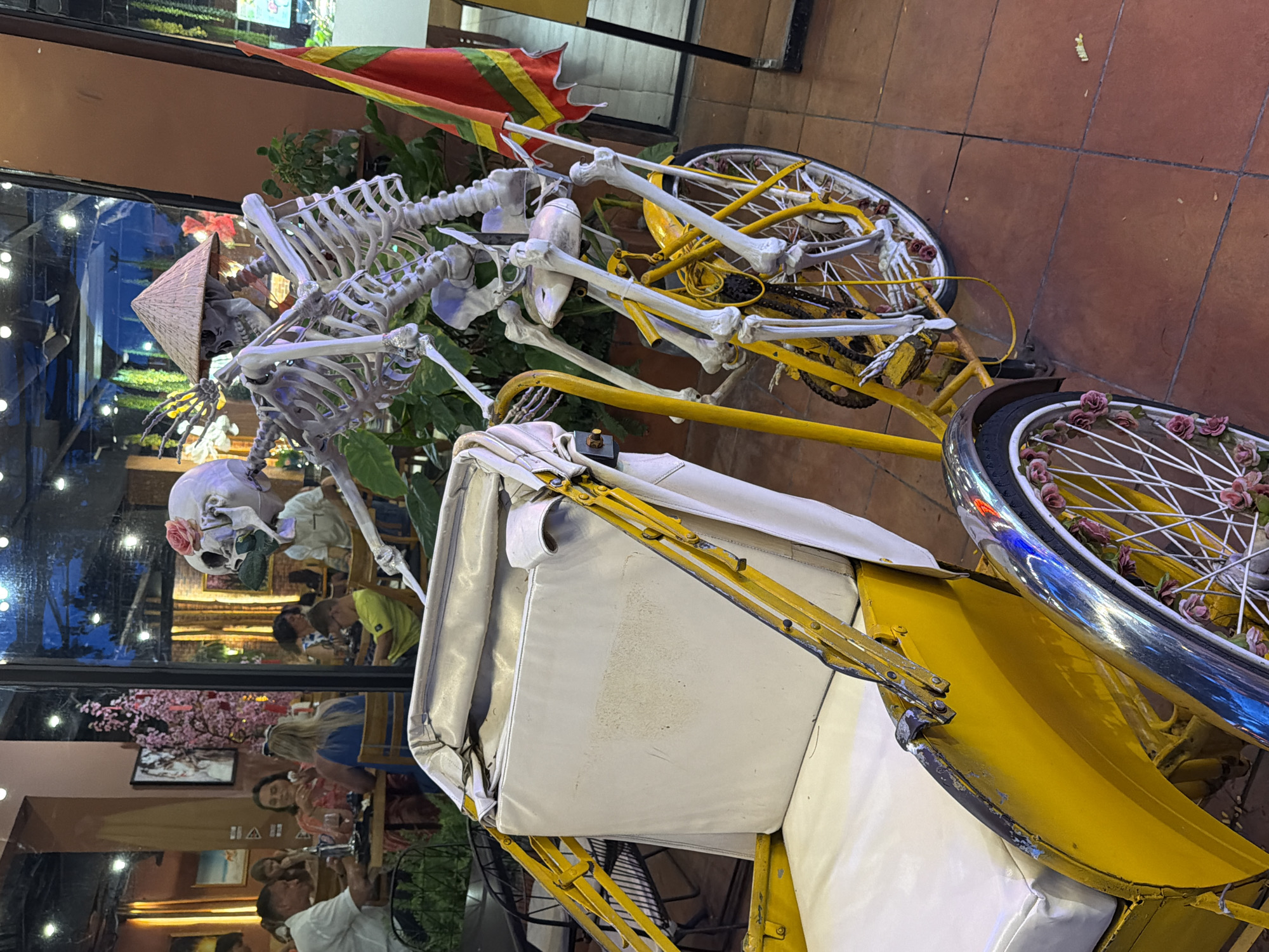 In Nha Trang, Vietnam, a yellow and white cyclopousse, a three-wheeled vehicle typically used as a taxi, is parked on a tiled floor in what appears to be an outdoor dining area of a restaurant. A skeleton figure, adorned with a conical hat and a pink flower on its skull, is positioned behind the driver's seat and appears to be operating the vehicle. The skeleton is leaning forward, with its bony arms positioned as if steering. A red and yellow striped flag is attached to the rear of the cyclopousse. In the background, through a glass window, several patrons are seated at tables, dining. The ambiance suggests a festive or thematic decoration within the restaurant, with foliage and decorative trees visible.