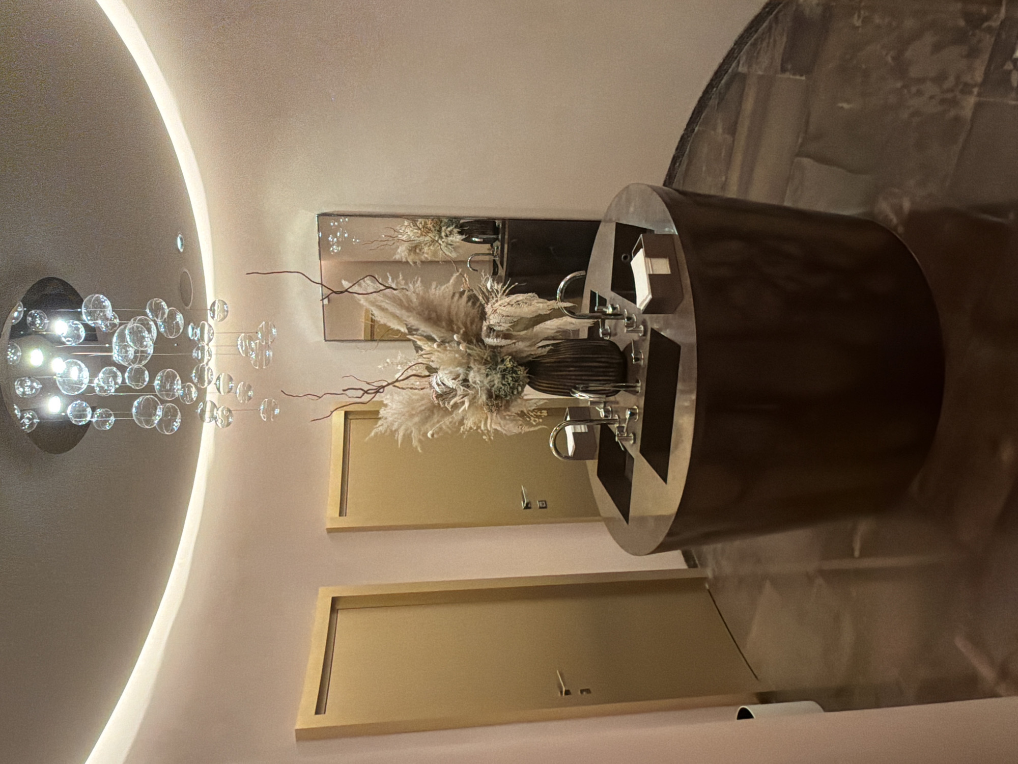 The image depicts a contemporary interior space, possibly a public restroom or a lobby area, with a modern aesthetic. The scene is dominated by soft, warm lighting, suggesting it is either nighttime or an indoor environment without natural light.
Prominently featured is a cylindrical, dark metallic vanity unit with a sleek countertop. On the countertop, a decorative floral arrangement of dried pampas grass and branches is displayed in a dark, ribbed vase. To the right of the floral arrangement, a faucet and sink are visible, suggesting a functional vanity area. Adjacent to the vanity, a mirror reflects the arrangement and the surrounding elements.
The background reveals two identical, rectangular doors with brushed gold frames, likely leading to individual stalls or rooms. Each door has a simple, modern handle. Above the doors, a mirrored panel reflects the upper portion of the room, including a portion of a curved ceiling.
To the left of the image, a unique chandelier composed of numerous clear glass spheres of varying sizes hangs from the curved ceiling. The chandelier is illuminated, casting a soft glow. The ceiling itself is a prominent architectural feature, with a smooth, curved surface that transitions into a wall. The overall ambiance is one of sophistication and modern design, with a focus on clean lines and subtle decorative elements. There is no visible text in the image. The location is identified as Saint Petersburg, Russia.