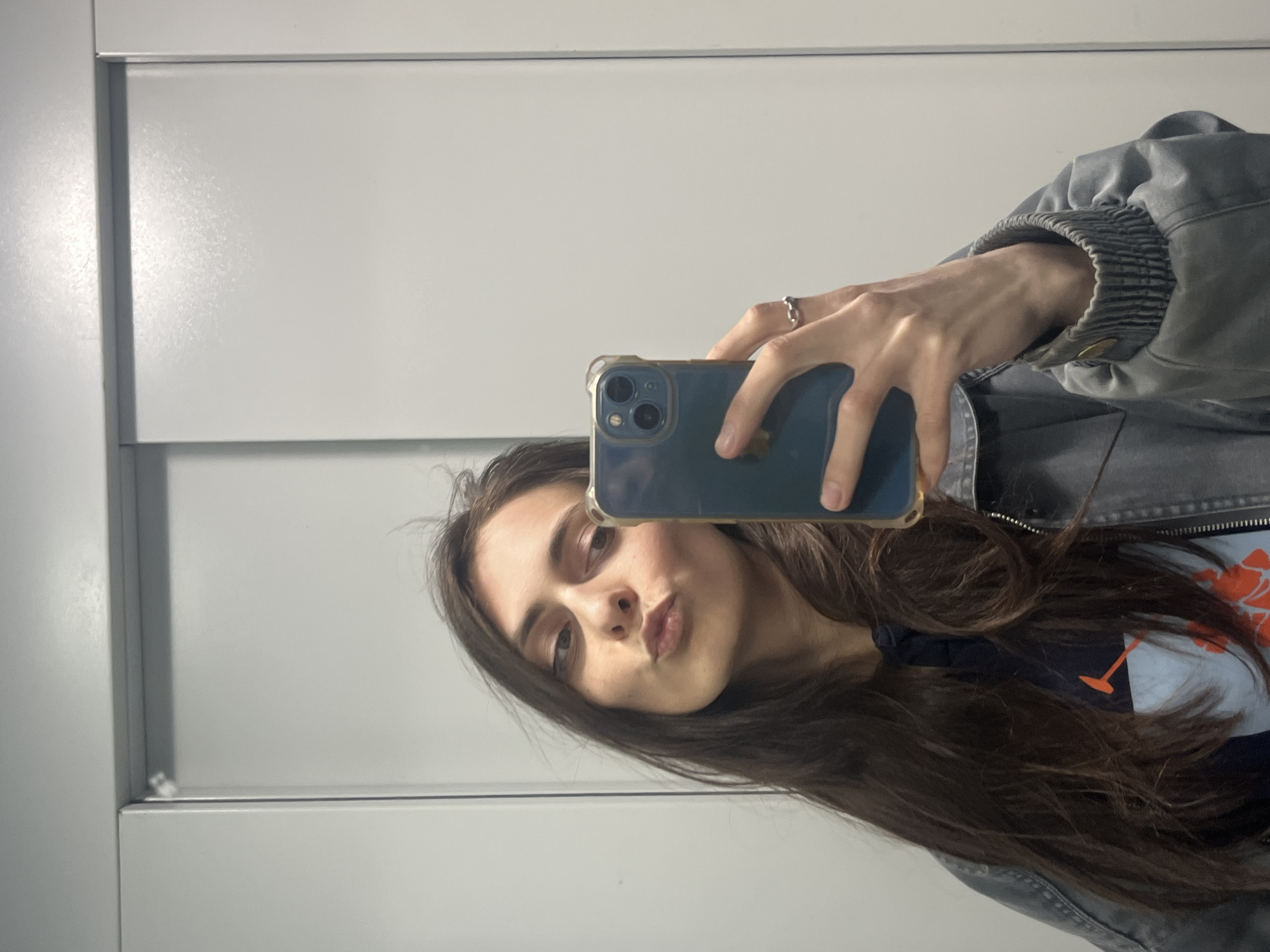 A young woman is taking a selfie in front of a white paneled wall, presumably a mirror or cabinet. She is holding a blue smartphone with a clear case in her right hand, obscuring part of her face. She has long, dark brown hair and is wearing a light blue shirt with a dark blue design underneath a gray denim jacket. A ring is visible on her right ring finger. The exact location in Krasnoyarsk, Russia, cannot be determined from the image.
