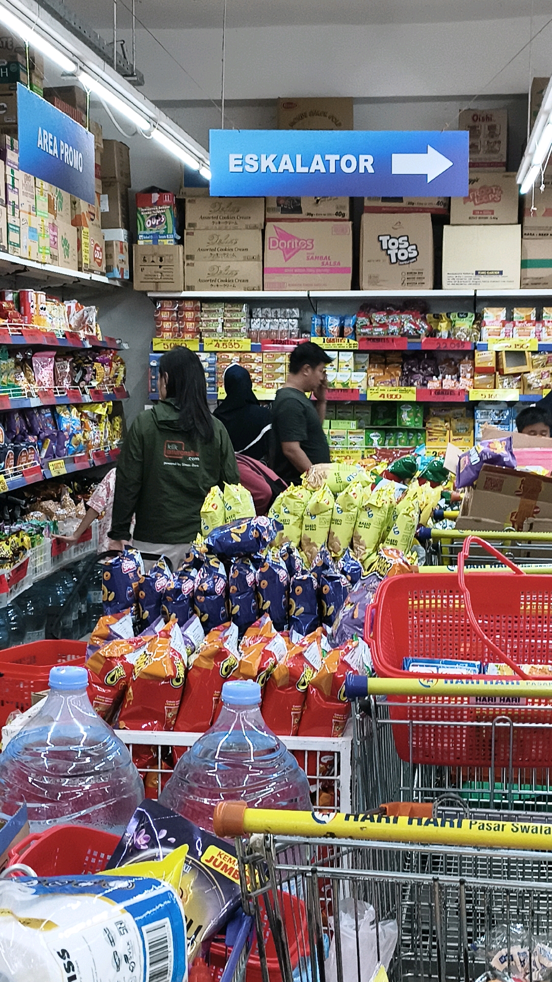 The image depicts the interior of a supermarket in West Jakarta, Indonesia. In the foreground, two large bottles of water are visible in shopping carts. Several red shopping baskets are also present. In the middle ground, shelves are stocked with various food products, including bags of snacks and chips. Three people are standing near the shelves, seemingly browsing or shopping. One person is wearing a green jacket and looking at the products, another is wearing a black headscarf, and a third is wearing a dark green shirt and appears to be speaking on a phone. Overhead, blue signs are hung, with one reading "AREA PROMO" and another "ESKALATOR" with an arrow pointing to the right. The overall environment is well-lit, suggesting an indoor commercial space.