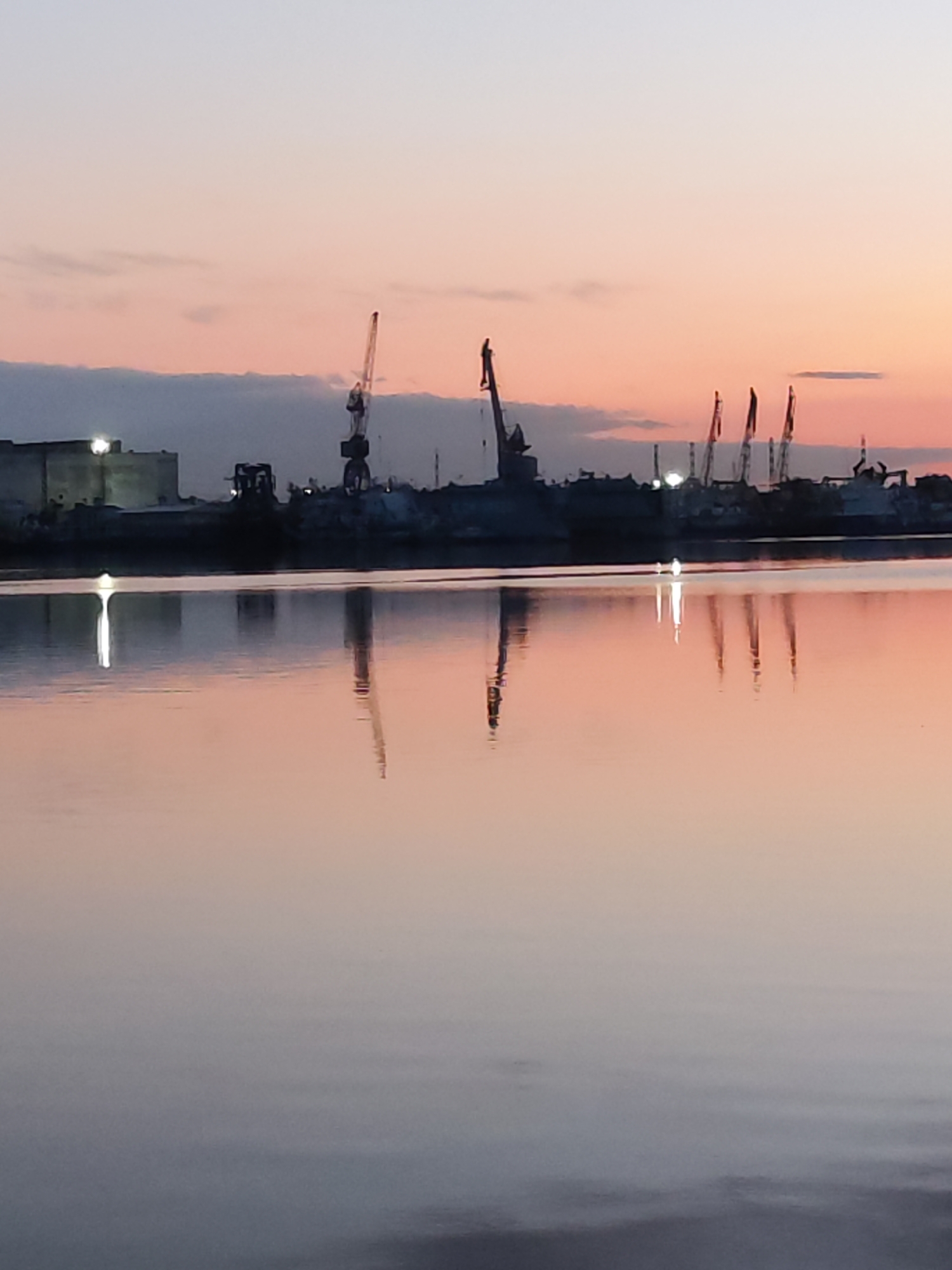 The image captures a serene view of a harbor or port area at dusk or dawn, most likely in Chaikovsky, Russia. Several industrial cranes dominate the skyline, their silhouettes reflected in the calm water below. The sky is painted with soft hues of pink and orange, creating a peaceful atmosphere. The water is mostly smooth with some small ripples. A few buildings or structures are visible on the shoreline, along with lights.
