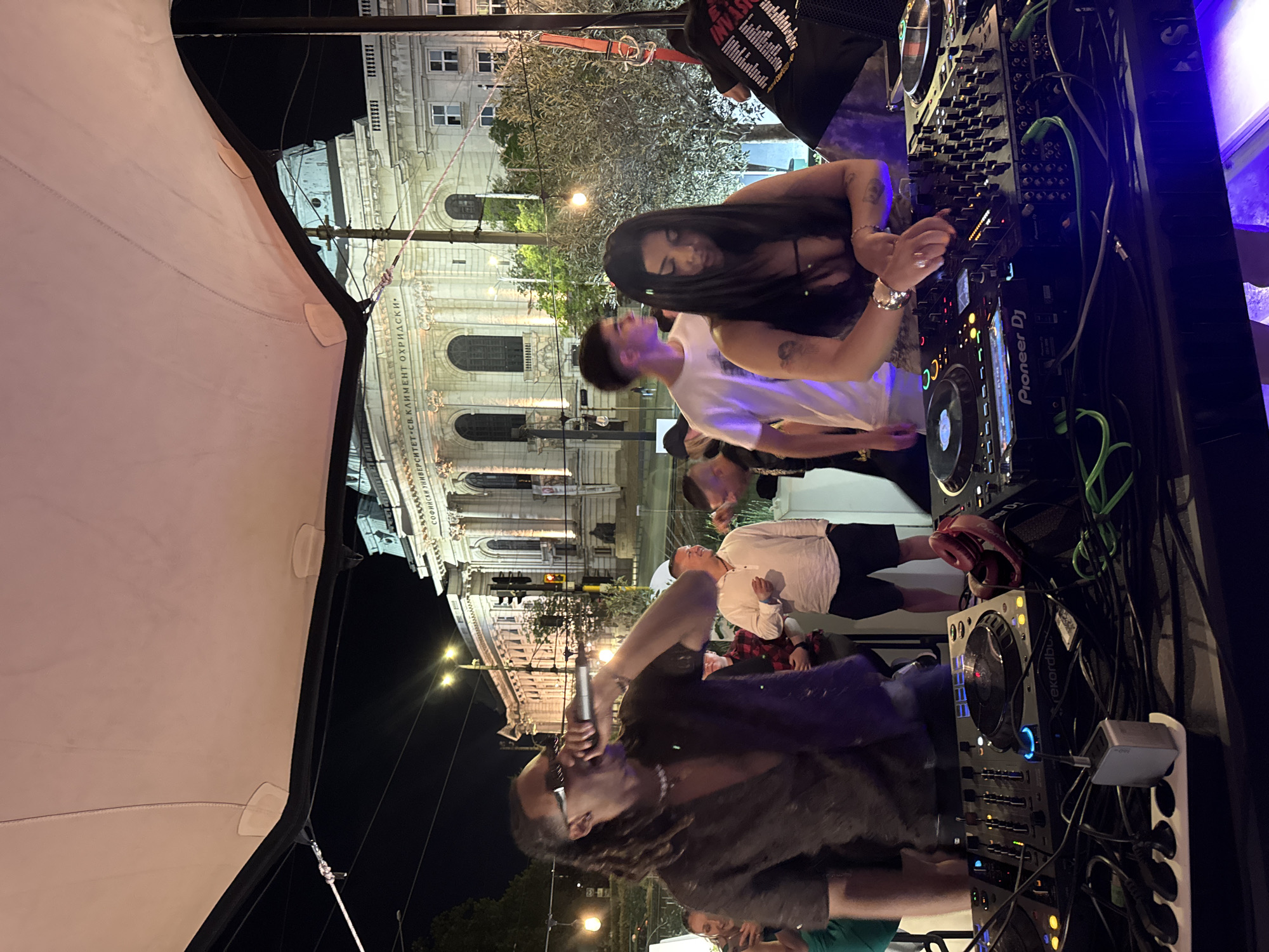The image depicts an outdoor event at night in Sofia, Bulgaria. A white tent-like structure is visible on the left. The facade of Sofia University "St. Kliment Ohridski" is in the background, illuminated by streetlights. A DJ booth is set up with electronic equipment, including Pioneer DJ decks and audio mixers. A woman with long dark hair is operating the equipment. A person is holding a microphone and speaking. Several people are present, including individuals wearing white shirts and casual attire. There are various cables and electronics.