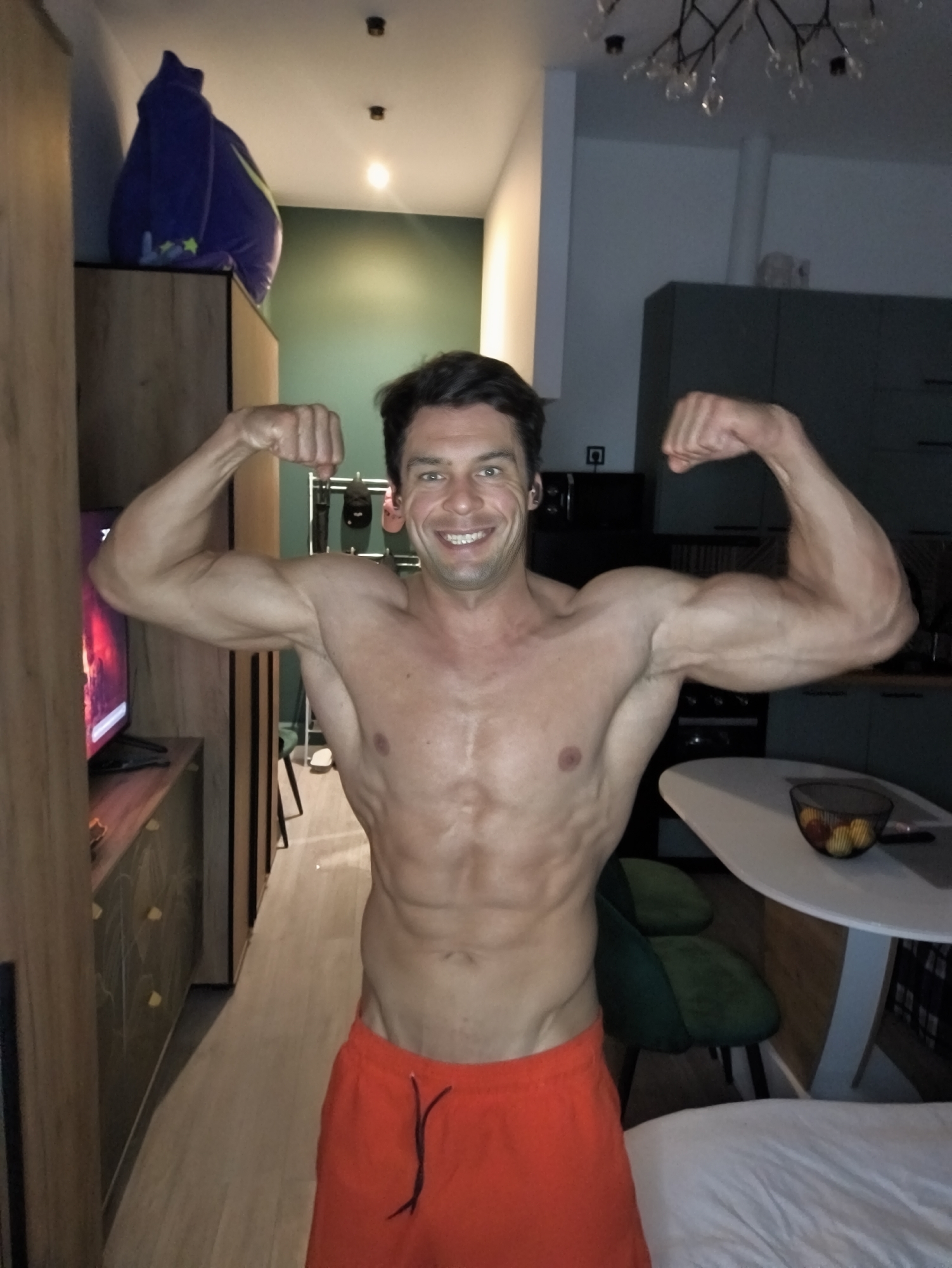A shirtless man is flexing his biceps in an interior setting. The man has short, dark hair and is wearing red shorts. His muscles are well-defined, particularly in his arms and abdomen. The background includes a television, a cupboard, and a beanbag chair on top of the cupboard. The interior design suggests a modern apartment. No specific evidence suggests that the location is in Moscow, Russia.