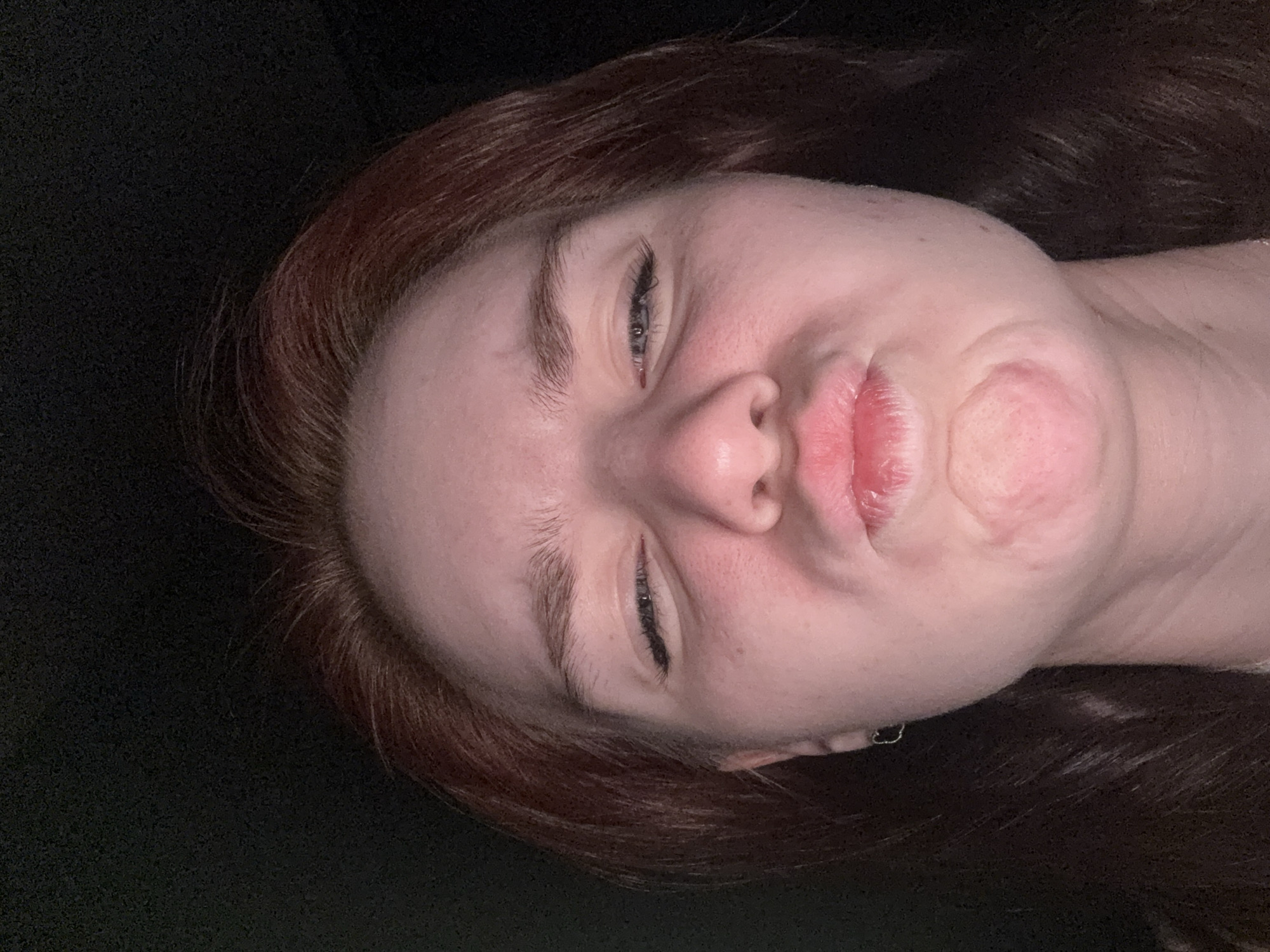 A close-up, angled selfie of a person with fair skin, red/brown hair, and blue eyes. The person's eyes are partially closed and their lips are puckered. A small earring is visible on the person's left ear. The background is dark and out of focus. The location is Unknown, Unknown.