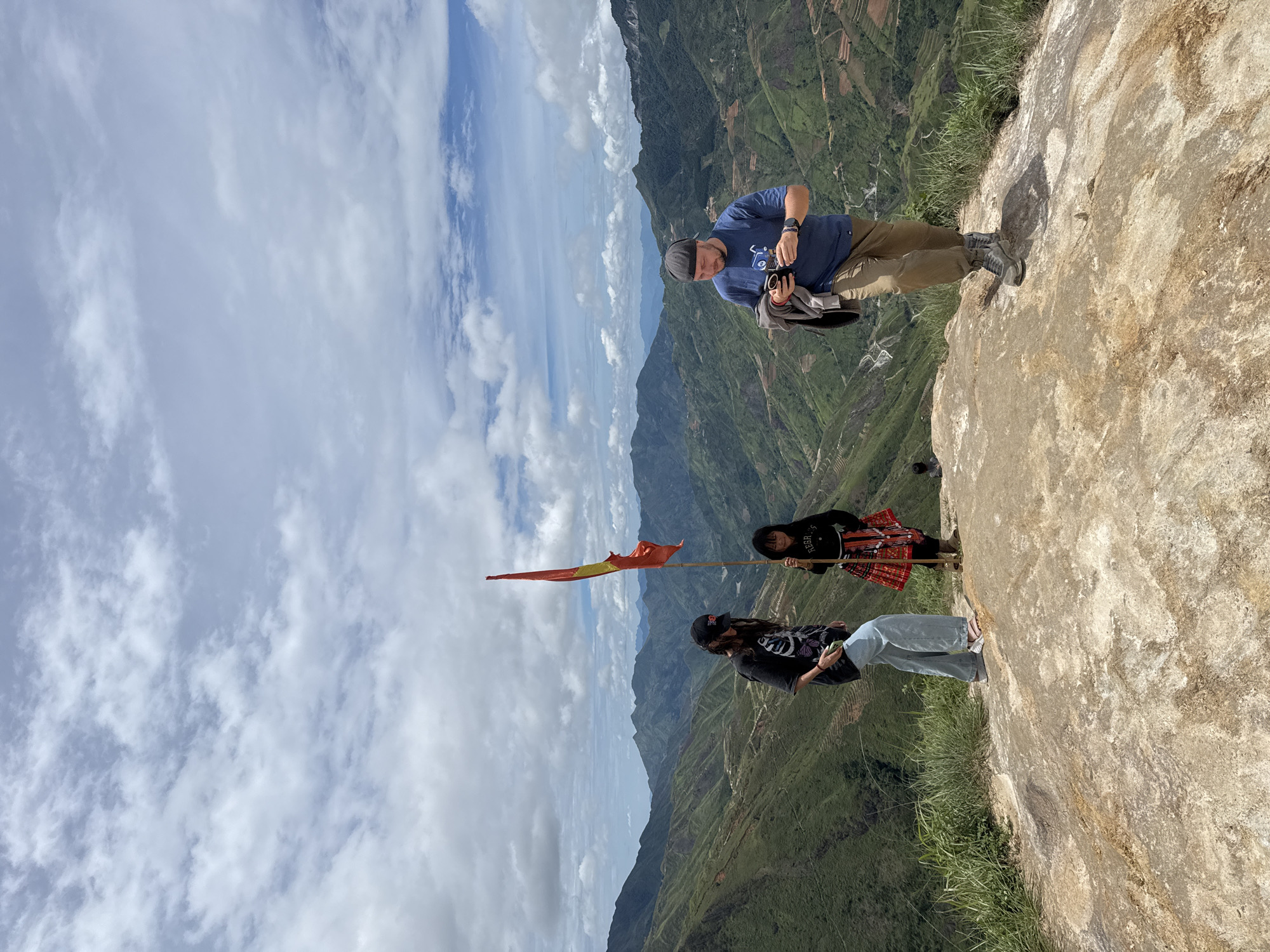 Three people are standing on a rocky outcrop overlooking a lush, mountainous landscape in Ta Xua, Vietnam. The sky is partly cloudy, with white clouds scattered across a pale blue expanse. In the foreground, a woman on the left, wearing a black t-shirt, jeans, and a baseball cap, holds a phone in her left hand. To her right, a young girl, dressed in a red and black traditional skirt and top, stands holding a bamboo pole with a red and yellow flag attached. On the far right, a man in a blue t-shirt, khaki pants, and a beanie holds a phone and a jacket. The background reveals rolling green hills with terraced fields and winding roads, indicative of the mountainous terrain of Ta Xua, a region known for its stunning natural beauty and hiking trails. The overall scene captures a moment of respite and enjoyment amidst a picturesque natural setting.