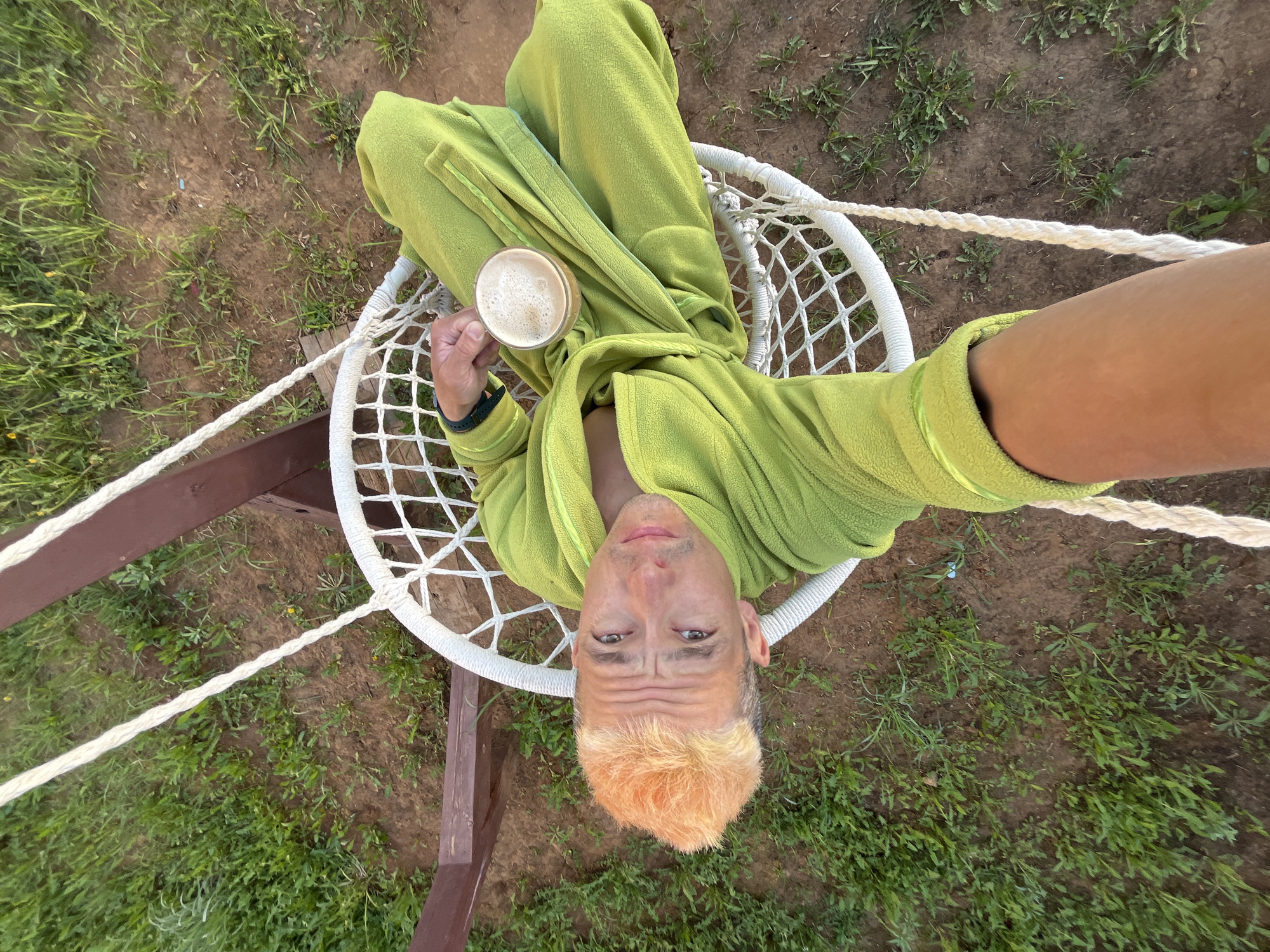 A man wearing a green robe is sitting in a white, webbed hammock chair in what appears to be an outdoor setting, possibly a yard or garden in Chaikovsky, Russia. He is holding a glass containing a beverage. The chair is suspended by ropes attached to a wooden frame. The ground around the frame is covered with dirt and some grass. The man's hair is dyed an orange color. The image seems to be taken as a selfie, with the man's arm extending towards the camera.
