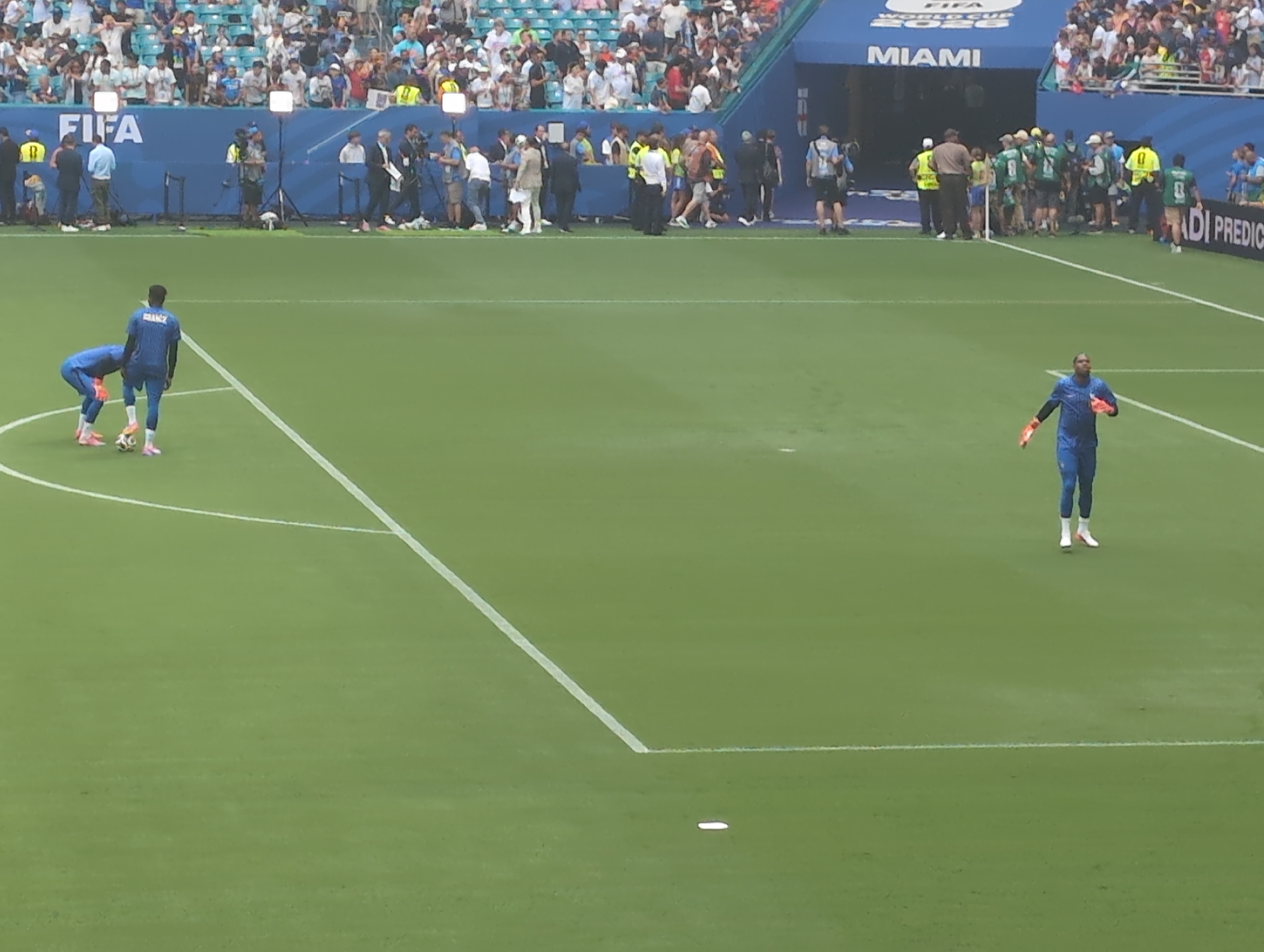 This image depicts a soccer field at Hard Rock Stadium in Miami Gardens, Florida, USA. Two soccer players in blue uniforms are visible on the field. One player, wearing a jersey with "FRANCE" across the back, is bending down to interact with a soccer ball. The other player is standing in a more upright position. In the background, a crowd of spectators is seated in the stands, and several individuals, including what appear to be security personnel and media members, are gathered near a tunnel entrance. The stadium's signage indicates it is a FIFA World Cup 2026 venue and specifically mentions "MIAMI." The field itself is marked with white lines, and the overall environment suggests a pre-game or practice session.