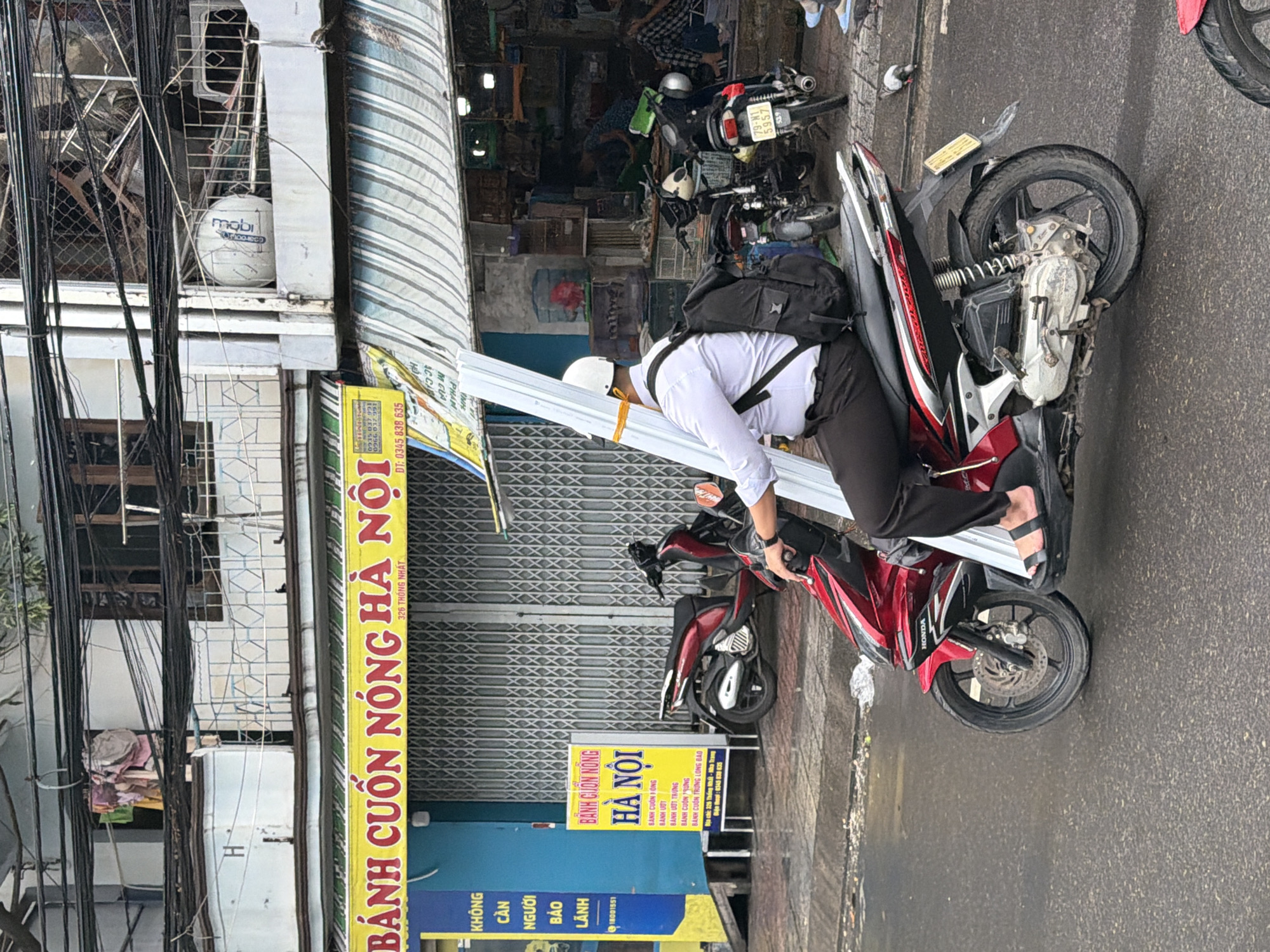A man wearing a white helmet and a white collared shirt with a black backpack is riding a red scooter down a wet street in Nha Trang, Vietnam. He is carrying long, white cylindrical objects strapped to his side, extending from the front to the back of the scooter. The scooter has a black seat and the brand name "HONDA" is visible on its side. The man is wearing black pants and black sandals. In the background, there are buildings with businesses, including a sign for "BÁNH CUỐN NÓNG HÀ NỘI" (Hanoi Hot Rolls). There are other scooters and people visible in the background. The street appears to be paved and wet, suggesting recent rain. The environment appears to be an urban street scene.
