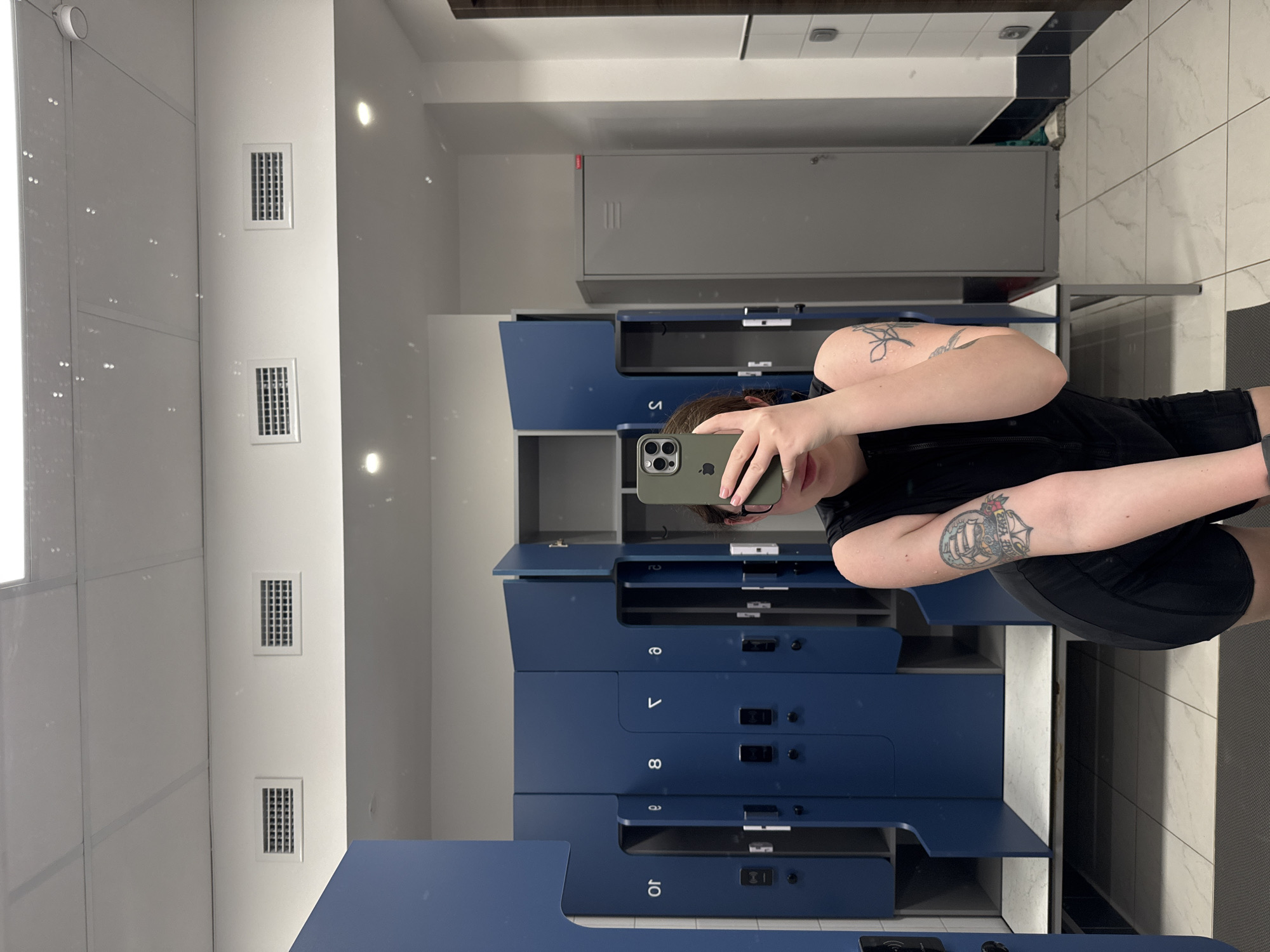 A person stands in a brightly lit locker room, reflected in a full-length mirror, holding up a cell phone to take a selfie. The person is wearing a black t-shirt and black shorts. Tattoos are visible on their arms. Behind the person are rows of blue lockers with silver handles and numbering from "5" to "10." A larger, gray locker is positioned above the blue lockers. The walls are white and feature rectangular ventilation grilles. The floor is tiled in a light grey color. A black, rectangular mat is on the floor near the mirror. The reflection reveals a portion of the ceiling, including a black trim piece. The location is potentially a locker room facility in a city similar to Chaikovsky, Russia, given the locker design and layout commonly found in such facilities, although there are no definitive identifiers within the image to confirm the location.