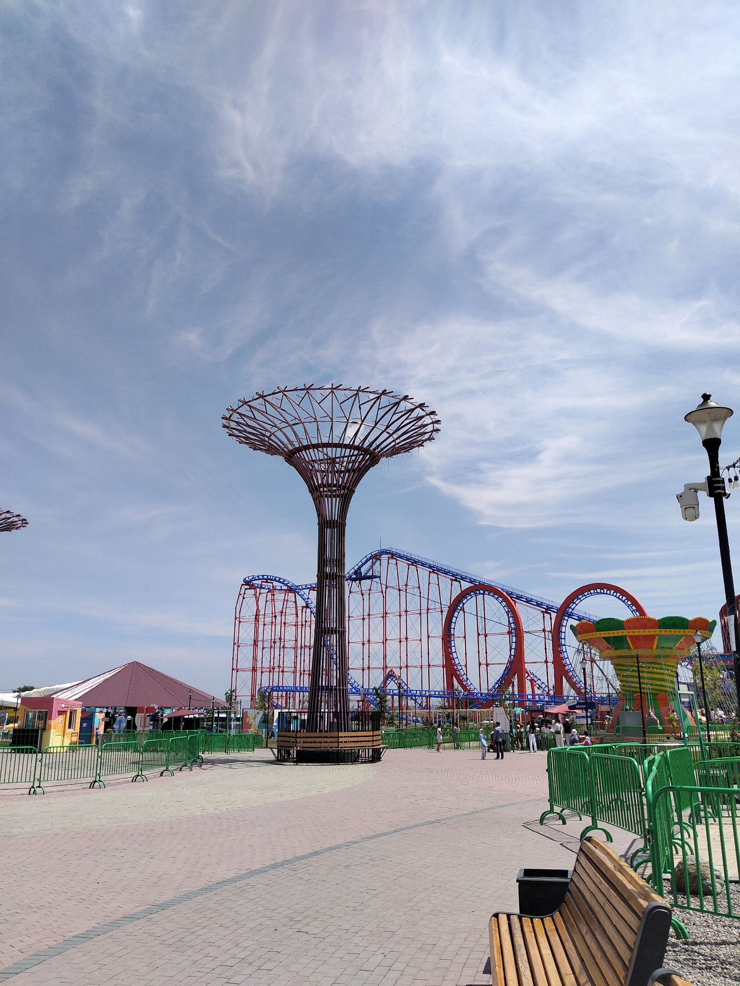 The image shows an amusement park with a roller coaster in the background and a tall, tree-like metal structure in the foreground. There are several people walking around the park, and various rides are visible, including a carousel. The ground is paved with red and gray bricks, and there are green fences throughout the park. The sky is blue with some white clouds. The amusement park is located in Bishkek City, Kyrgyzstan.
