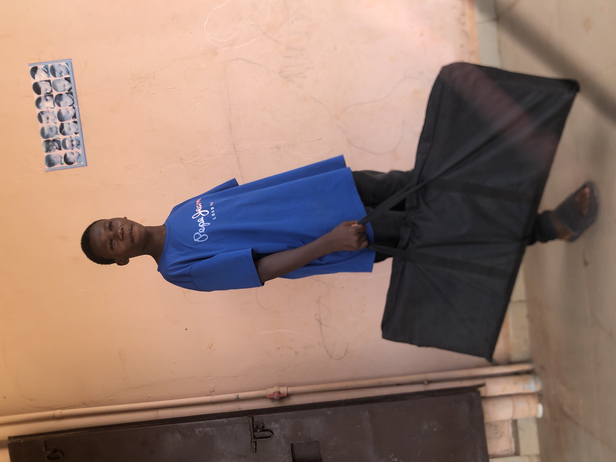 An African male stands in front of a light-peach exterior wall in Nkwelle Ezunaka, Nigeria. He wears a blue t-shirt with a logo on the left breast and black pants. He is holding a large black rectangular bag with straps. The wall has a photo strip of multiple images of a baby, as well as some white marks or scrawls. A brown door or panel sits at the base of the wall with pipes running along it. The ground is either tiled or concrete. The man wears one sandal on his right foot.