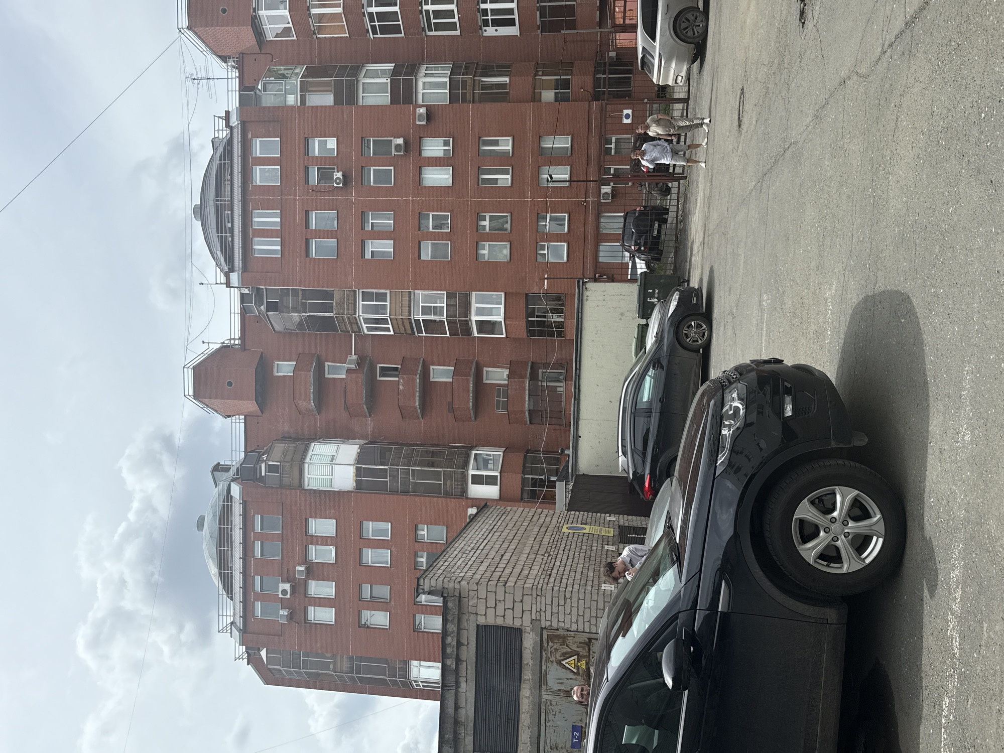 The image captures a residential building in Perm, Russia, characterized by its red brick facade and a distinctive domed roof section. The building appears to be a multi-story apartment complex with numerous windows and balconies. In the foreground, a dark colored SUV is parked on an asphalt surface that shows signs of wear and tear. To the left of the building, a smaller, utilitarian structure with a corrugated metal roof is visible. Two men are standing near a metal gate in the middle ground, with several other vehicles parked behind them and to the right. The sky is overcast with scattered clouds.