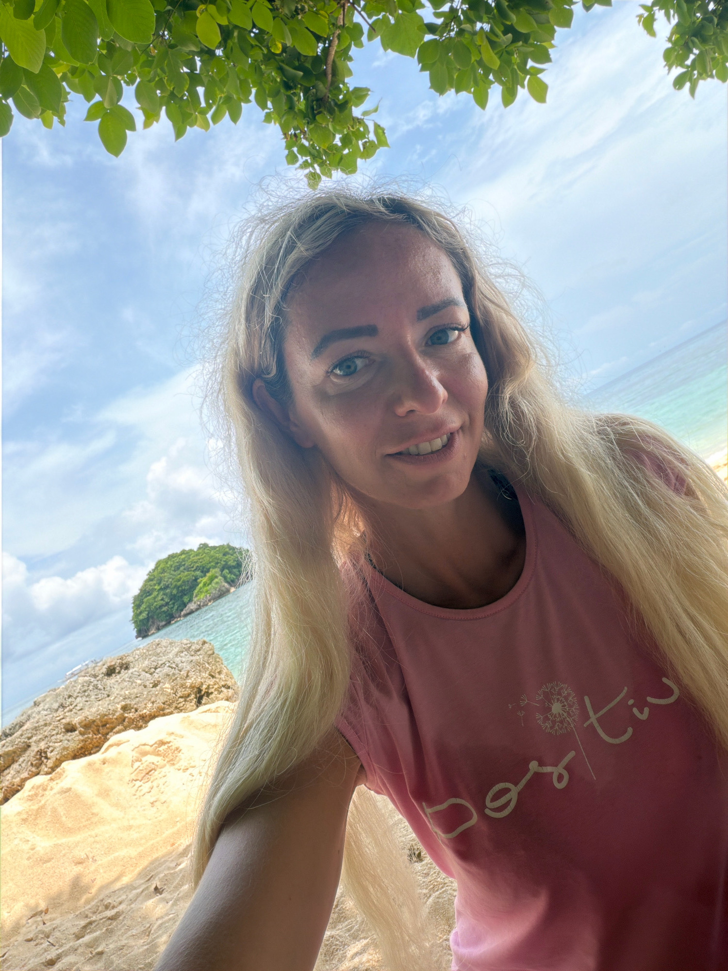 A woman with blonde hair and blue eyes takes a selfie on a beach in Malay, Philippines. She is wearing a pink t-shirt with a dandelion graphic and the word "positiv" printed on it. Behind her, lush green trees frame the sky, and the turquoise waters of the ocean meet a sandy shore. A small, verdant island rises from the sea in the distance. The sun is shining, casting a warm glow on the scene.