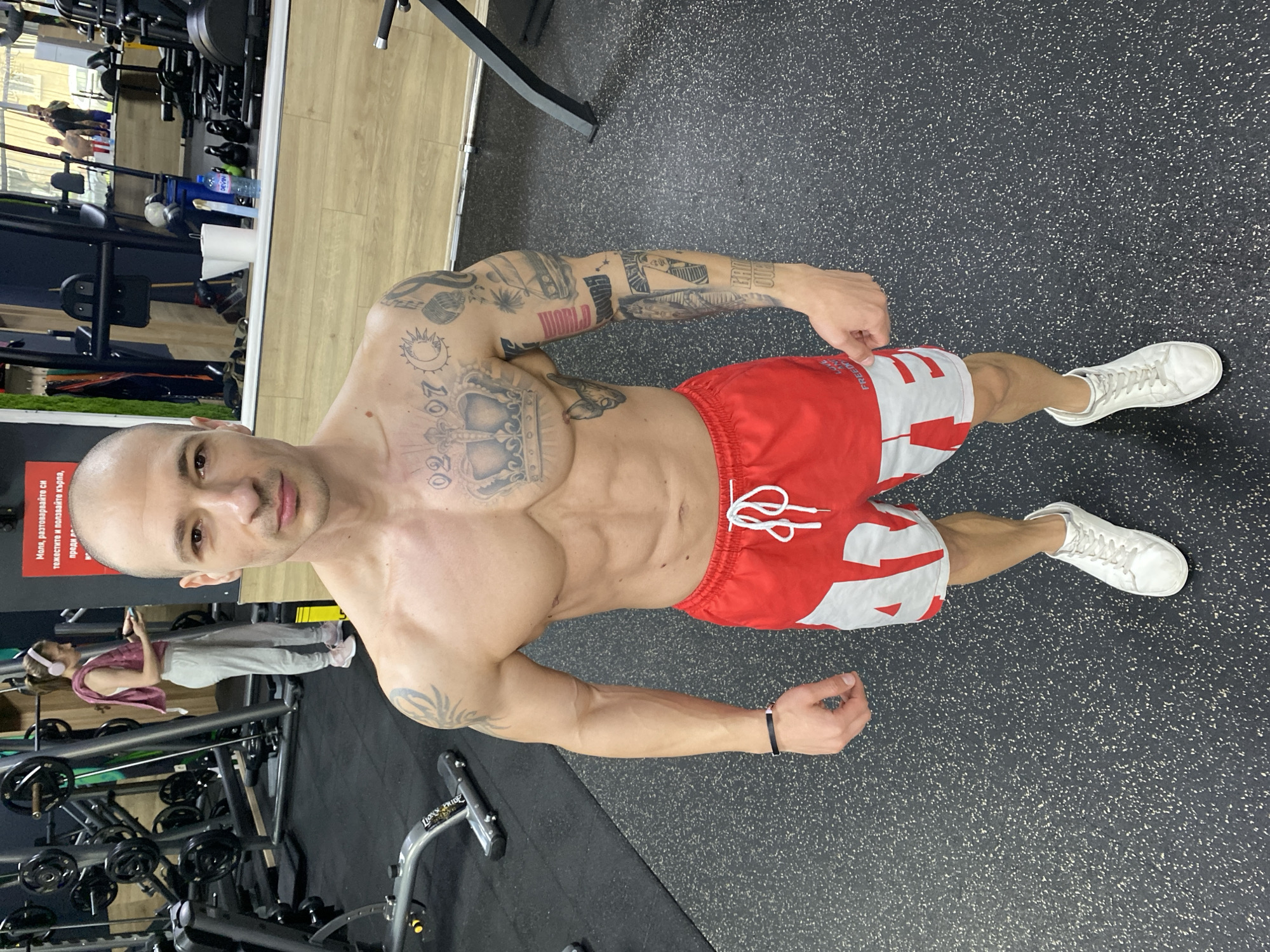 A muscular, bald man stands in a gym, facing the camera. He has numerous tattoos on his chest and arms. He is wearing red and white shorts and white sneakers. The gym contains exercise equipment, and there is a woman in the background on a treadmill. A sign in Bulgarian is partially visible on the left. The gym is located in Pleven, Bulgaria, as the sign contains Bulgarian text and one of the tattoos on the man's arm reads "Pleven".