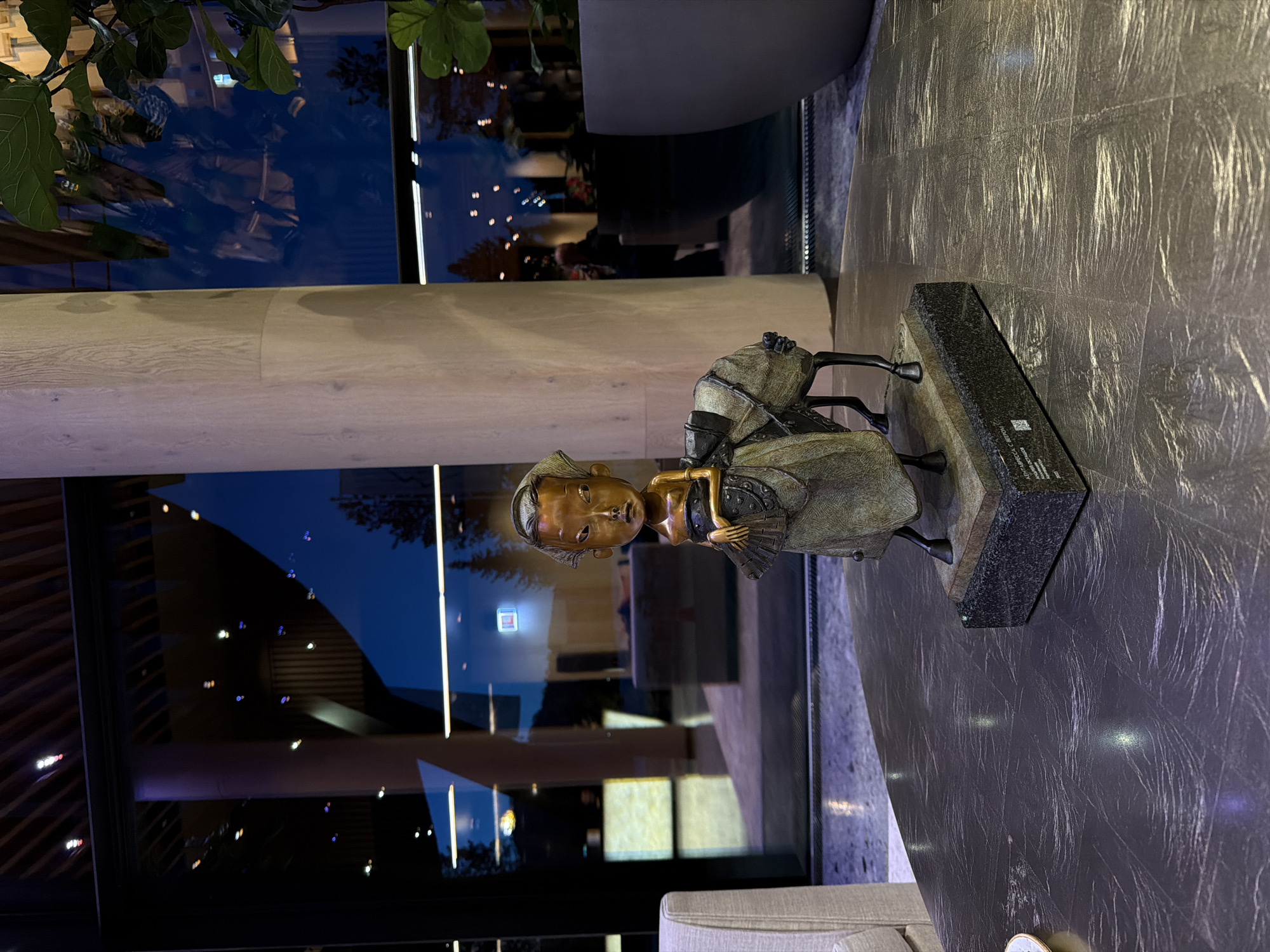 A medium shot depicts a bronze statue of a seated figure with a fan, placed on a granite plinth on a grey-toned table. The statue is positioned near a large window reflecting an interior setting. A structural column is to the left of the window, and part of a plant is visible in the upper-left corner. The surrounding environment suggests an upscale interior, but there are no specific indicators to confirm the location in Озёрное, Russia.