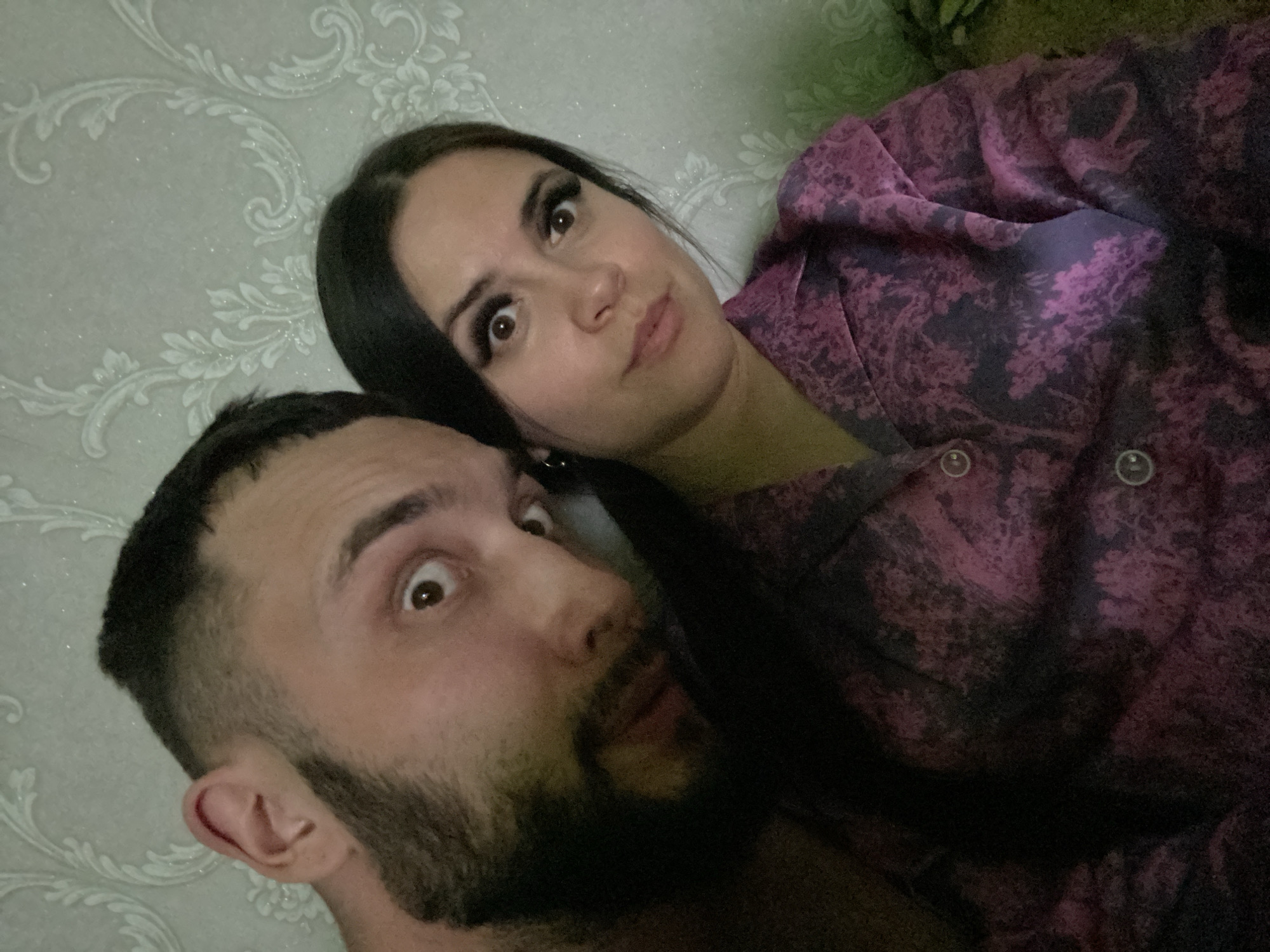 A man and a woman are captured in a close-up photograph. The man, positioned in the foreground, has dark hair and a beard. His eyes are wide with a startled expression. The woman, located behind him, also has dark hair and is wearing a dark pink patterned top. Her facial expression is inquisitive. The background features a wallpaper with a repeating floral pattern. The location is within the city of Chaikovsky, Russia.