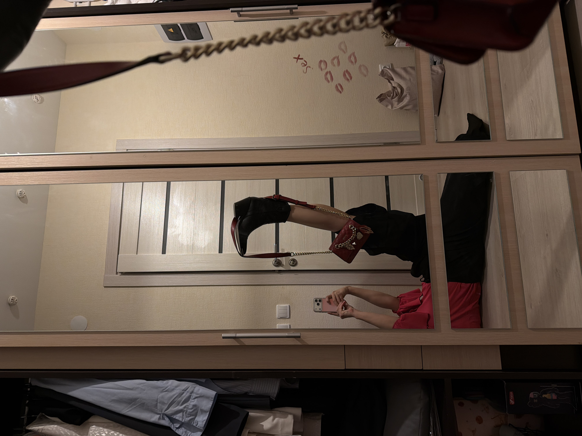The image depicts a person taking a photo in a room with mirrored wardrobe doors. The room has light-colored walls. In the reflection, a person is visible taking a picture with a mobile phone. The person is wearing red shorts and is reflected in the wardrobe mirror. The person also has a black boot on their leg and a red purse hanging from their leg.
The wardrobe contains clothes. There are also decorative elements on the wall in the reflection of the mirror, including the word "sex" and lipstick marks.
There is nothing visible in the image that indicates it is in Izhevsk, Russia.