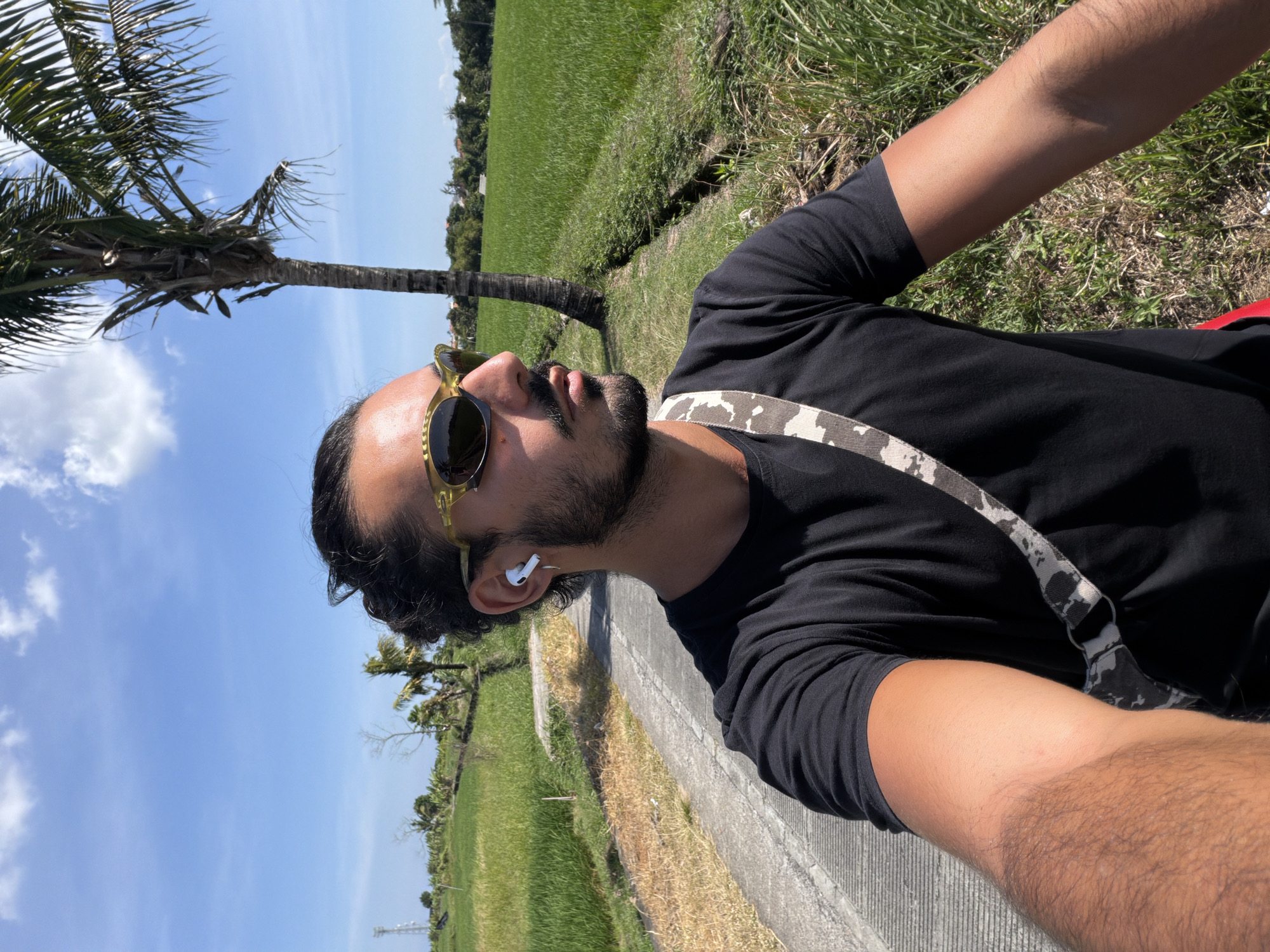 A medium shot captures a man in a black t-shirt with a camouflage strap, wearing green-tinted sunglasses and white wireless earbuds, as he looks towards the right. He has a beard and his hair is tied back in a bun. The man is standing next to a paved path that runs through a bright green rice paddy field under a clear blue sky with scattered clouds. A tall palm tree is visible to the right of the man. The scene appears to be in North Kuta, Indonesia, a region known for its rice fields and tropical vegetation.
