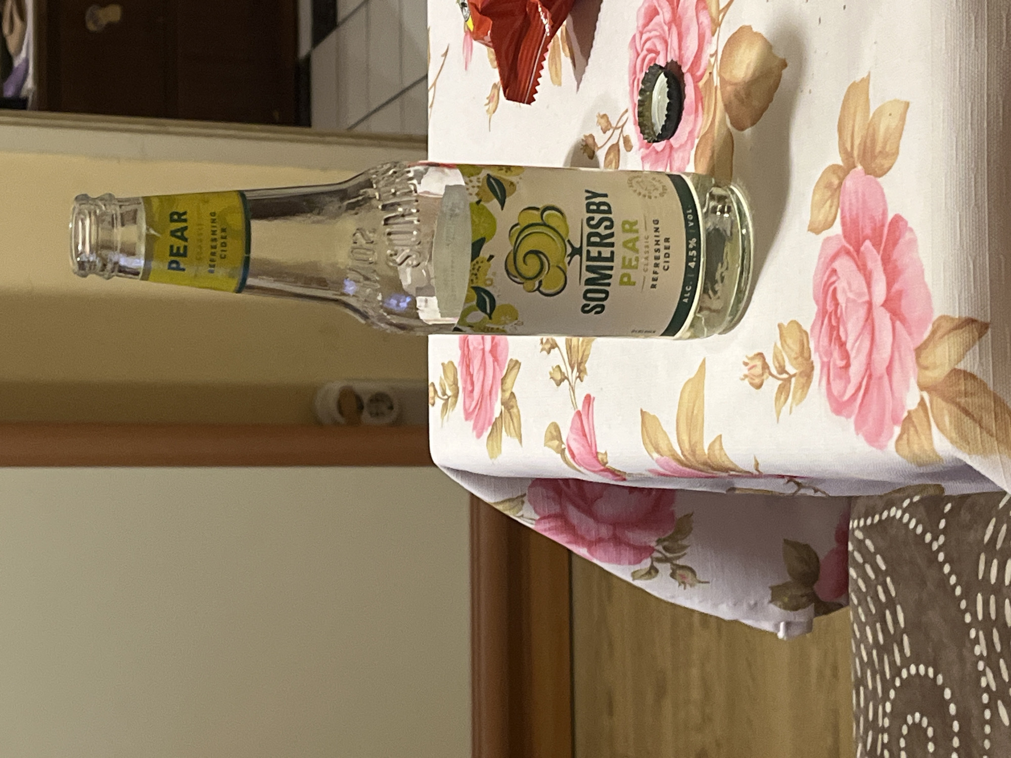 A clear glass bottle of Somersby Pear cider stands upright on a table covered with a white tablecloth adorned with pink roses and green leaves. The bottle has a yellow label with "PEAR" prominently displayed in black text, along with "CLASSIC REFRESHING CIDER" and the alcohol percentage "ALC. 4.5% VOL.". A silver bottle cap lies uncapped next to the bottle. To the right of the bottle, a corner of a red bag of snacks is visible. The background shows a wall with an electrical outlet and a dark wooden dresser. The scene is indoors, likely in a home, and the lighting suggests it is daytime. The image does not provide any specific information that can be used to determine its location in the city of Plovdiv, Bulgaria.