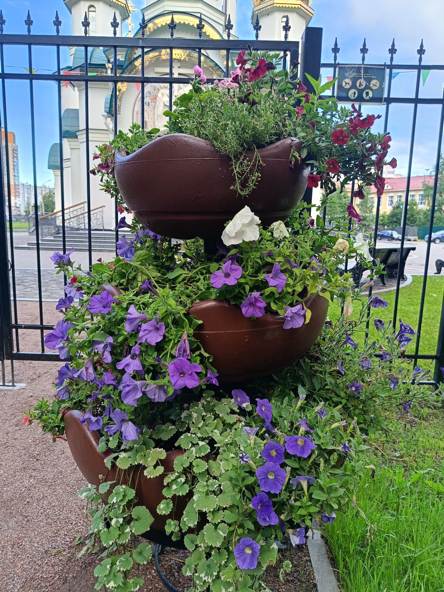 A multi-tiered planter filled with flowers is positioned outdoors, in front of a black metal fence. The planter contains a variety of flowers, including purple petunias and some red and white blossoms. Behind the fence, a large white building with ornate gold trim and spires is visible, suggesting it might be a church or a historical building. The sky is blue and partly cloudy. The foreground features gravel and grass. The scene appears to be in a public area, likely a park or near a significant landmark within Saint Petersburg, Russia, given the architectural style of the building.