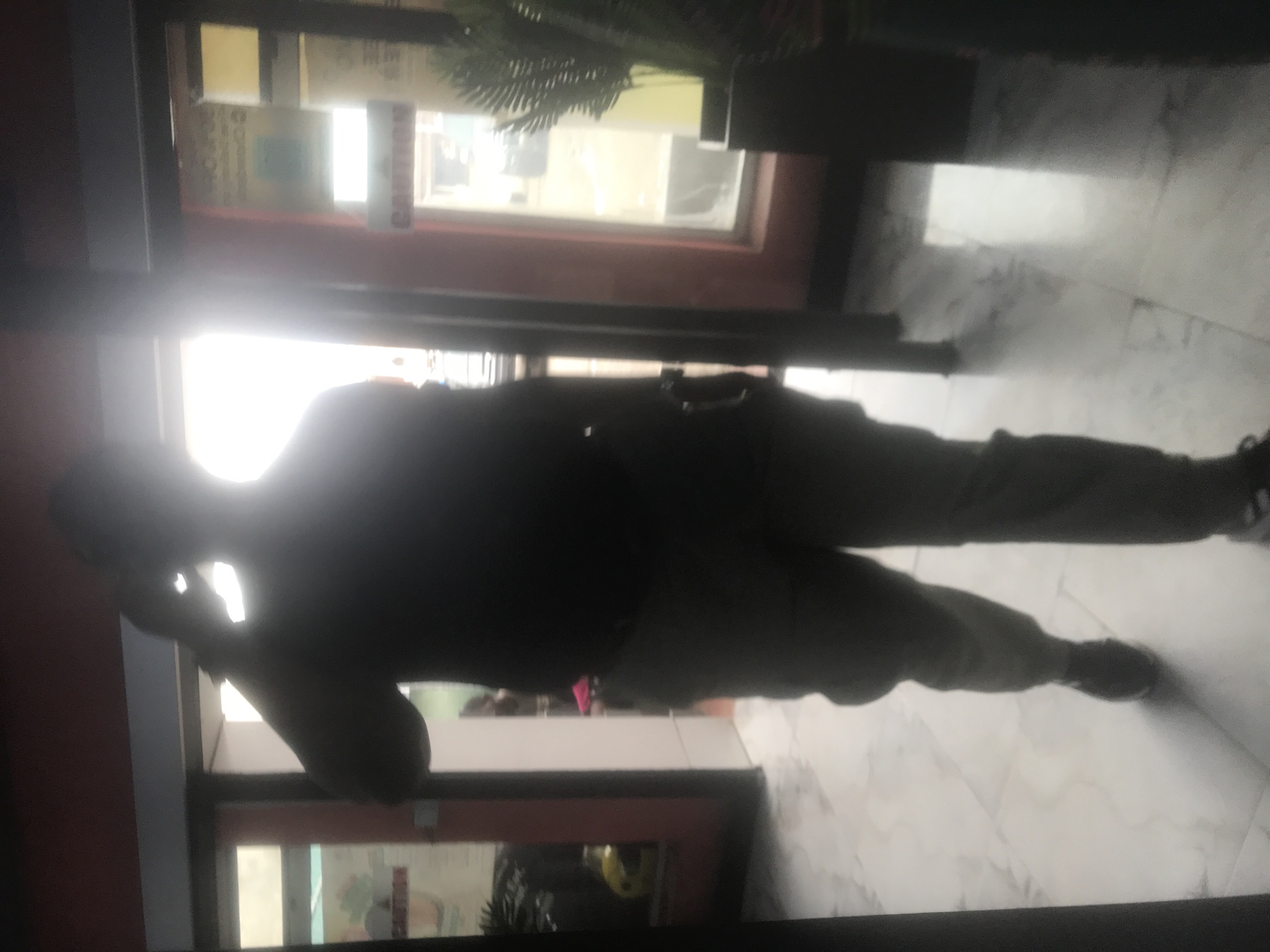 The image, captured in landscape orientation and appearing to be taken indoors, features a silhouette of a person standing with their back to the camera. The person is wearing dark-colored pants and a dark top, with their left arm raised and bent, appearing to be holding something to their head. A bright light source is positioned behind the person, creating a strong silhouette and obscuring finer details of their appearance and the surroundings.
The background suggests an interior space with large windows or glass doors. Through these, a blurry view of an outdoor or another indoor area is visible, with hints of sunlight and possibly some greenery. The flooring appears to be tiled with a light color. The overall lighting is dramatic due to the backlighting, emphasizing the dark outline of the person.
No specific activity or event can be definitively identified due to the silhouette and the quality of the image. The time of day is suggested to be daytime by the bright light, but the indoor setting makes it difficult to be certain. There is no discernible weather information. Location cues are limited to an interior space, possibly a shop, office, or residential building with large windows. No text is clearly visible in the image. The emotion conveyed is neutral due to the lack of discernible facial features or body language beyond the pose.
