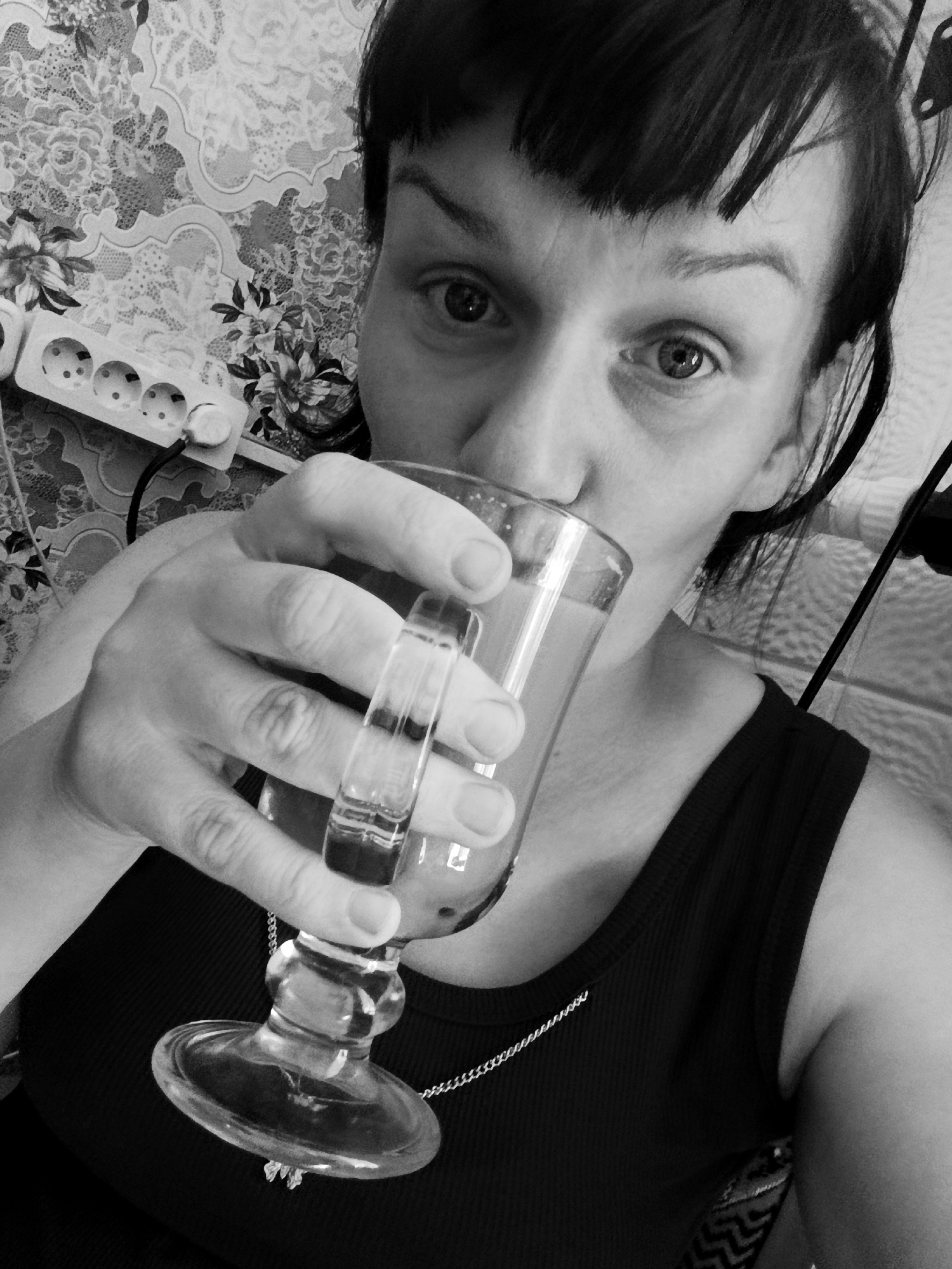 A monochrome, close-up shot shows a woman drinking from a clear glass with a wide base. The woman is positioned at an angle, facing the camera. She has dark hair with bangs, and her eyes are open wide, looking directly at the camera. She's wearing a dark tank top and a thin silver chain necklace. Her hand holds the glass. Behind her, there is a wall with a floral pattern, and to the upper left, a white power strip with multiple outlets is visible. The location, Фоки, Russia is not discernible from the image.
