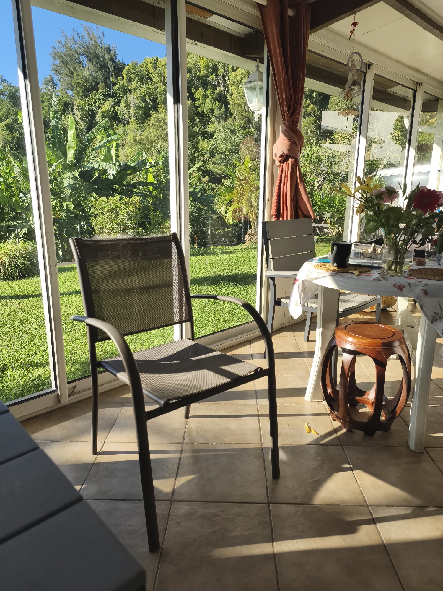 The image depicts a sunlit sunroom overlooking a lush, green outdoor landscape. In the foreground, a dark metal chair with a mesh seat and back is positioned on a tiled floor. To the right, a white table is partially visible, covered with a patterned tablecloth. On the table, a vase holds a bouquet of flowers, including red and pink blooms, alongside a dark mug. A decorative wooden stool with an open, curved design sits beside the table. Sunlight streams into the room, casting distinct shadows on the floor. Through the glass walls of the sunroom, a vibrant scene of tropical vegetation, including banana trees and dense foliage, is visible under a clear blue sky. There are no people in the image. The setting is likely a residential sunroom in Saint-Denis, France, characterized by its connection to a garden environment.