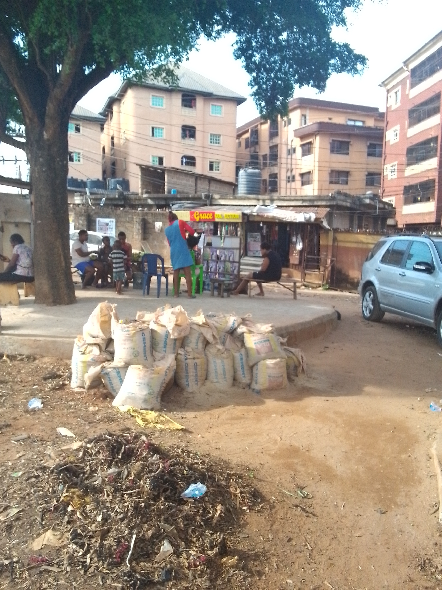 The image depicts a scene in Onitsha, Nigeria, showing a cluster of buildings, several people, and a streetscape with litter and bags of cement.
In the foreground, there is a pile of dry leaves and debris on the ground. Behind this, a collection of about a dozen off-white bags labeled "CEMENT" are stacked together.
In the midground, several people are visible. Some are sitting around a small makeshift table or platform, while others appear to be standing and interacting with a shop. The shop has a sign with "Grace" written on it. A woman in a red shirt and blue dress is standing near the shop.
In the background, there are multiple multistory buildings of similar design and color, indicating an urban residential area. The lighting suggests it is daytime. A silver SUV is parked on the right side of the frame. The scene has a bustling feel, characteristic of a busy street-side location in Onitsha.