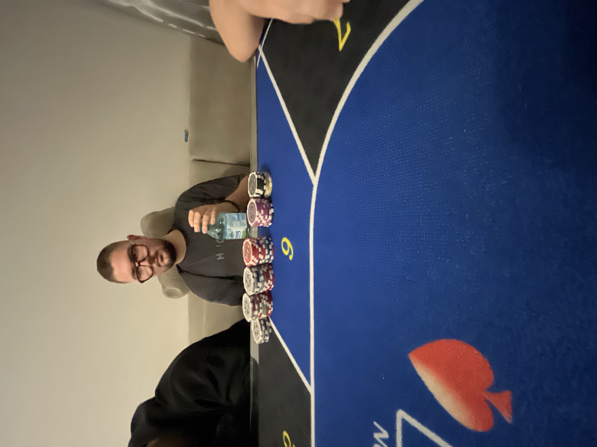 A man with glasses, wearing a gray t-shirt, is sitting at a poker table with stacks of chips in front of him. He is holding a water bottle. Another person in a black shirt is partially visible to the left. The poker table is blue with suit markings. A couch is visible in the background. There is no information in the image to determine that the location is Burgas, Bulgaria.