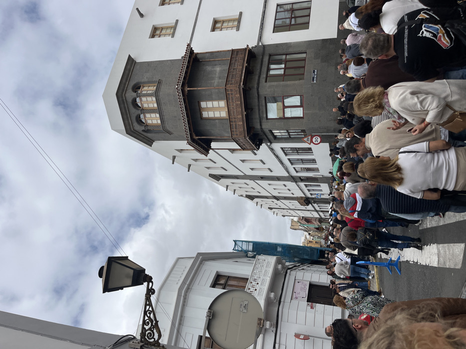 The image shows a street scene in the city of Las Palmas de Gran Canaria, Spain, located at coordinates 28.098 latitude and -15.416 longitude. The scene is crowded with people walking in both directions. The people are of various ages and ethnicities. Some are wearing casual clothing, while others are wearing more formal attire. Many are carrying bags and purses. There are buildings on both sides of the street. The buildings are mostly white with some gray accents. There is a streetlight on the left side of the street. The street is paved and has crosswalks painted on it. The sky is cloudy and the sun is not visible. The image was taken on a weekday afternoon.