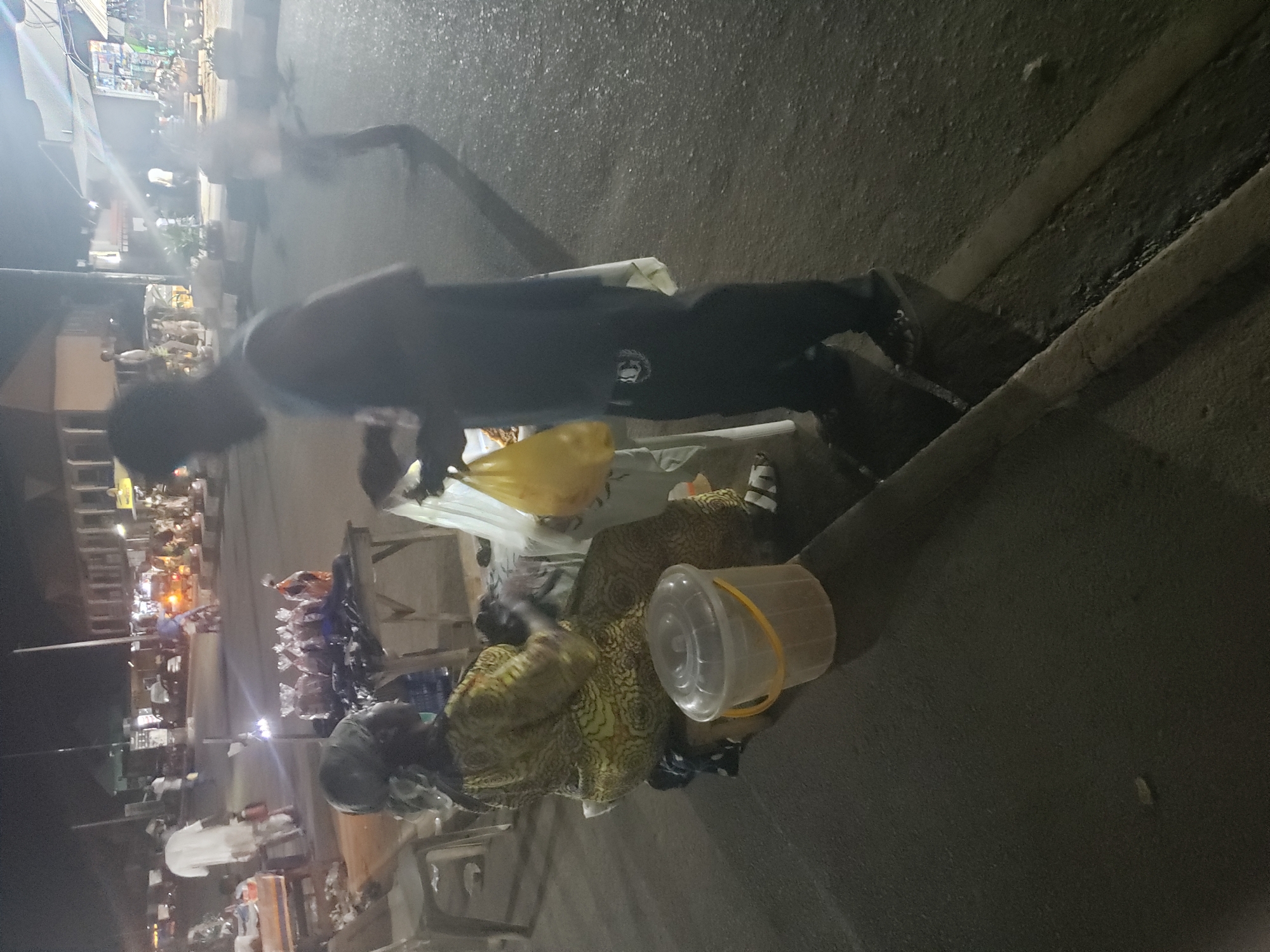 The image presents a vibrant night street scene, likely a bustling market or commercial area in Ibadan, Nigeria. The ambient light is dim, indicating it is evening or night, with artificial lights from stalls and shops illuminating the activity.
In the foreground to the right, a person is seated on the ground, wearing a patterned garment in shades of yellow and green, suggestive of traditional African fabric. Beside them is a clear plastic bucket with a yellow handle, containing plastic bags.
In the mid-ground, a standing person, presumably male, is visible. He is dressed in a dark long-sleeved top with a white circular crest or emblem on the chest, and dark trousers. He appears to be positioned near a market stall or table, which holds various items, including a prominent yellow plastic bag.
The background shows a lively street lined with numerous market stalls or small shops, which are lit up to display their merchandise. Several other blurred figures, likely vendors and customers, are present, contributing to the busy atmosphere. The ground is paved, resembling a street or pathway, with curbs or drainage channels running along the side.
The scene captures typical evening market activity where goods are traded. While specific emotions are not discernible due to the low light and general distance, the environment appears active and routine. No explicit text is clearly legible, beyond the indistinct logo on the standing person's clothing.