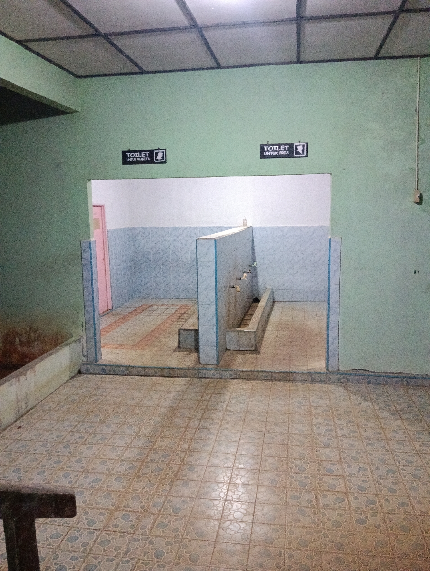 The image depicts an interior view of a public restroom facility, likely in Pematang Jaya, Indonesia, with separate sections for men and women. The walls are painted a light green, and the ceiling is made of acoustic tiles with a grid pattern. Two doorways lead into the restroom areas, each marked with a sign indicating "TOILET" and either a female or male silhouette.
The women's restroom area, visible on the left, appears to have a pink door. The men's restroom area on the right is open and shows a row of faucets mounted on a tiled wall. In front of the faucets is a long, shallow trough, possibly a urinal. The floor is tiled with a pattern of light brown and gray squares. The overall condition suggests a functional but not modern facility, with some visible wear and tear. A dark, partially visible object, possibly furniture or a fixture, is in the foreground on the left.