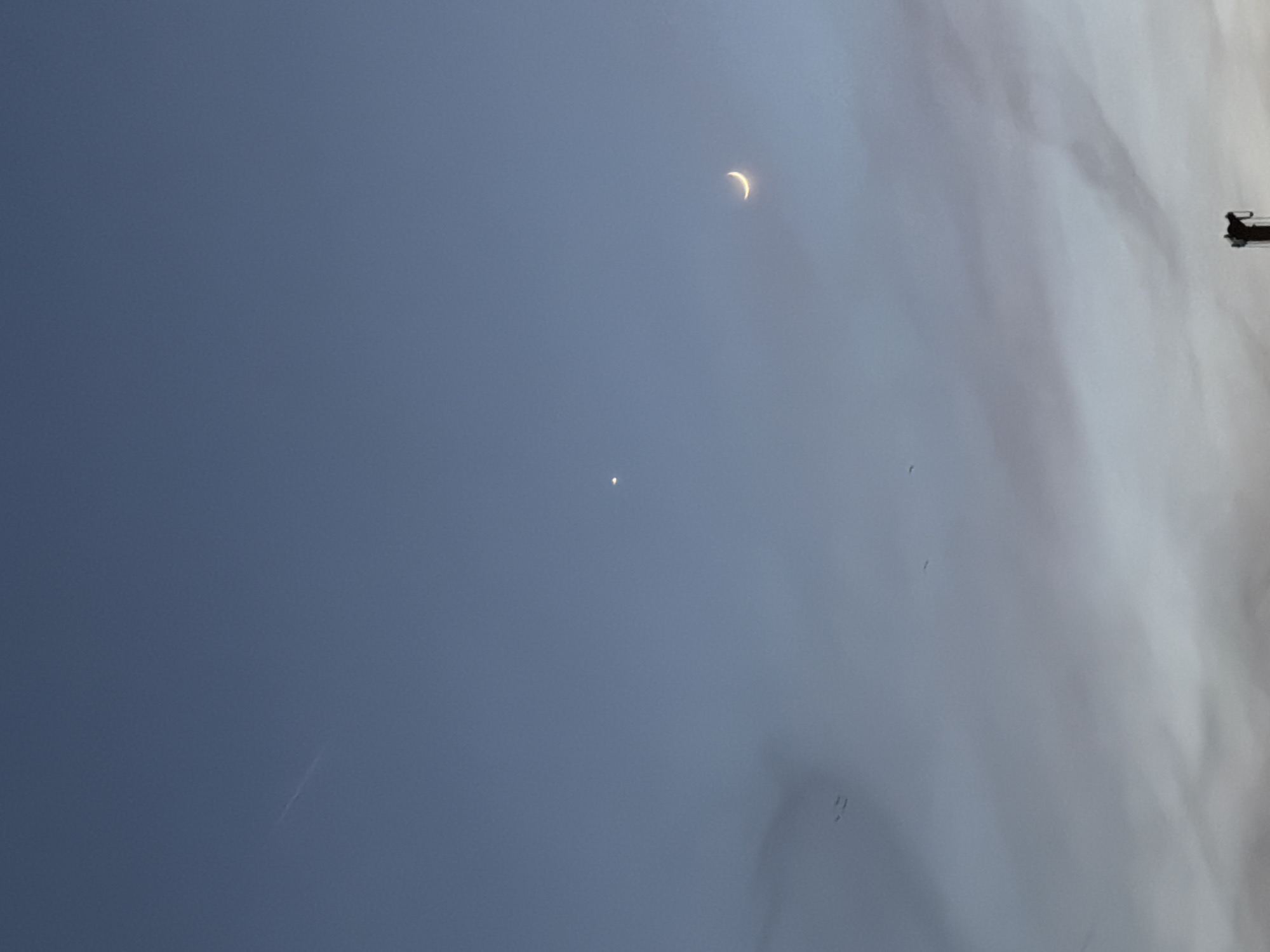 A dusk or dawn sky showing a thin crescent moon and a bright celestial object (likely Venus or Jupiter) below it. Clouds are present in the lower right. A vapor trail from an airplane is visible in the lower left. A utility pole is visible on the right side of the sky. It is not possible to determine if the location is Belgrade, Serbia from the visual information available in this image.