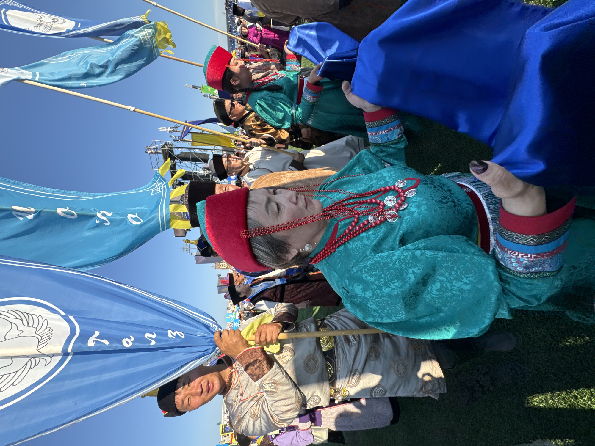 The image captures a vibrant scene at an outdoor event in Ulan-Ude, Russia. The main subjects are individuals dressed in traditional Mongolian attire, prominently featuring colorful robes, ornate headwear, and intricate jewelry. One woman in the foreground wears a striking turquoise robe adorned with a patterned fabric and a red hat. She is accessorized with a heavy, multi-layered red beaded necklace featuring silver decorative elements. Another person, also in traditional dress, is visible in the lower part of the frame, holding a blue flag with white lettering and a circular emblem depicting a bird.
Several flags are visible throughout the image, held aloft on wooden poles. These flags are in various shades of blue and yellow, some bearing script and symbols, contributing to the festive atmosphere. The people appear to be seated or standing in a line, suggesting participation in a procession or ceremony. Their expressions are largely neutral or slightly smiling, hinting at a sense of pride and cultural engagement.
The setting appears to be a grassy field, with hints of infrastructure like scaffolding and distant crowds suggesting a large public gathering or festival. The clear blue sky indicates fair weather and likely daytime, possibly mid-morning or afternoon, given the angle of the sunlight casting shadows. The overall mood is one of cultural celebration and community. No specific text is clearly legible beyond what appears to be Mongolian script on the blue flag.