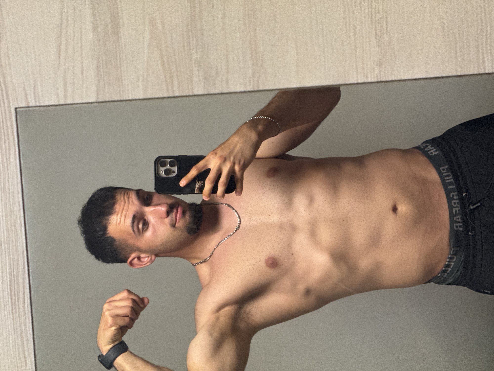 The image shows a shirtless man in a bathroom, taking a mirror selfie. He is flexing his bicep and holding a smartphone in his right hand. He is wearing dark-colored shorts with a visible logo on the waistband. The bathroom has light-colored wood-paneled walls and a grey wall. The man has short dark hair and a goatee. He is wearing a silver chain around his neck and a silver bracelet on his left wrist. The lighting is soft and even. There is no visible indication of the specific location within the city of Shumen, Bulgaria.