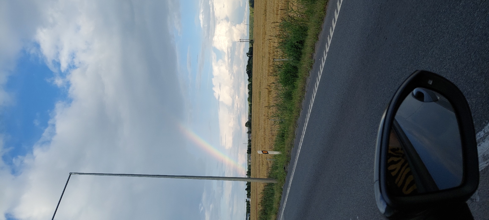 The image captures a scenic view from a car on a road, likely in Bernatoniai, Lithuania. A segment of a rainbow arcs across a partly cloudy sky, with a muted sun attempting to break through the clouds. The road in the foreground is asphalt with a white broken line, and to the right of the road is a grassy verge followed by a field of dry, golden crops, possibly wheat or barley. In the distance, a treeline borders the field, and beyond that, several structures and power lines are visible, suggesting a rural settlement. A solitary lamppost stands tall on the left side of the road. A side-view mirror of the car is prominently featured in the bottom left corner of the image, reflecting the sky and a yellow and black striped object, possibly clothing or an accessory. The overall atmosphere is serene and evocative of a recent or impending rain shower.