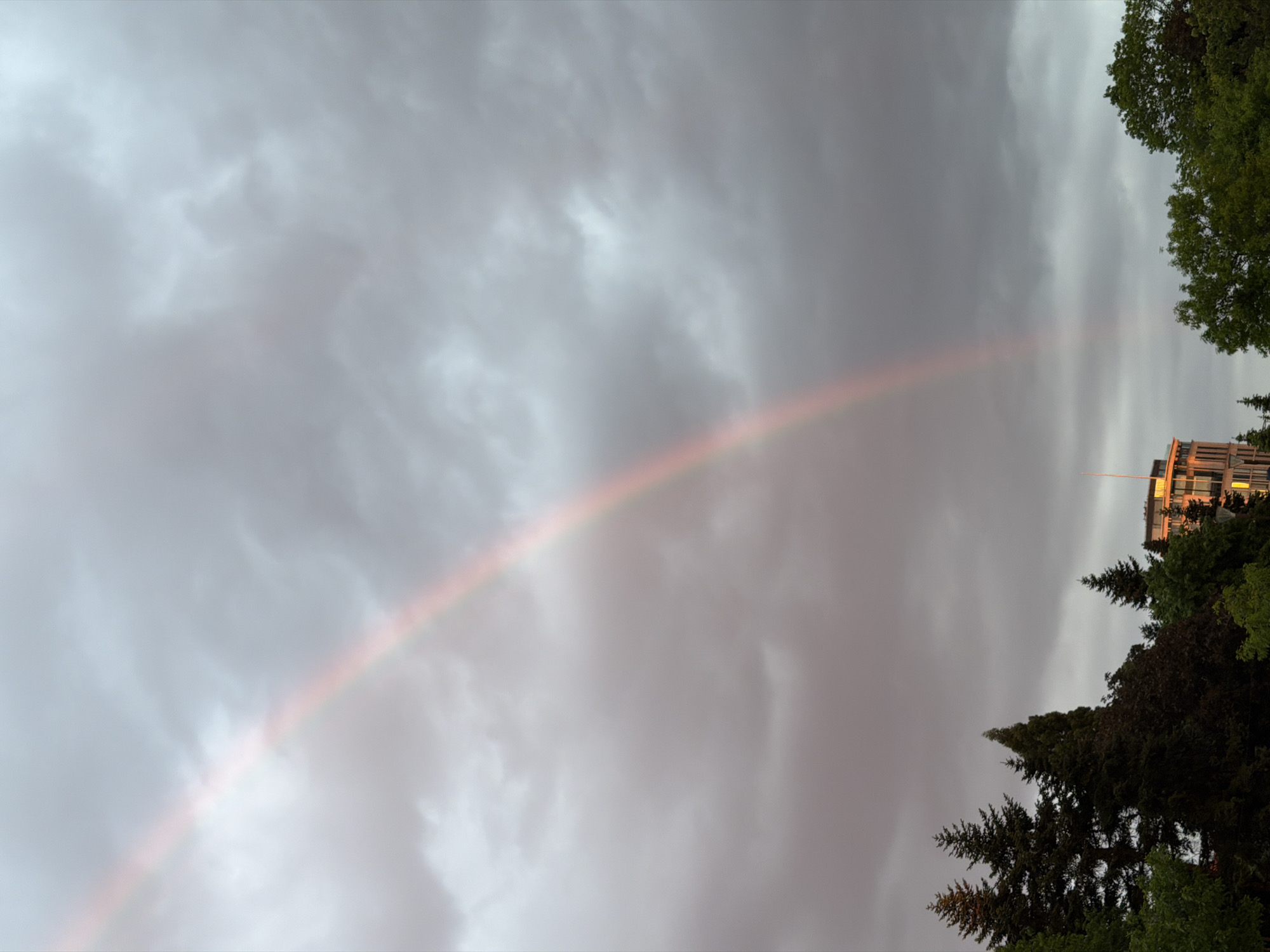 A medium shot captures a faint rainbow arching across a cloudy sky, partially obscured by gray and white clouds. The right side of the image shows a building with illuminated windows and a radio antenna, partially concealed by coniferous and deciduous trees. The trees occupy a significant portion of the lower right and upper right corners of the frame. This image could possibly be in Sofia, Bulgaria.