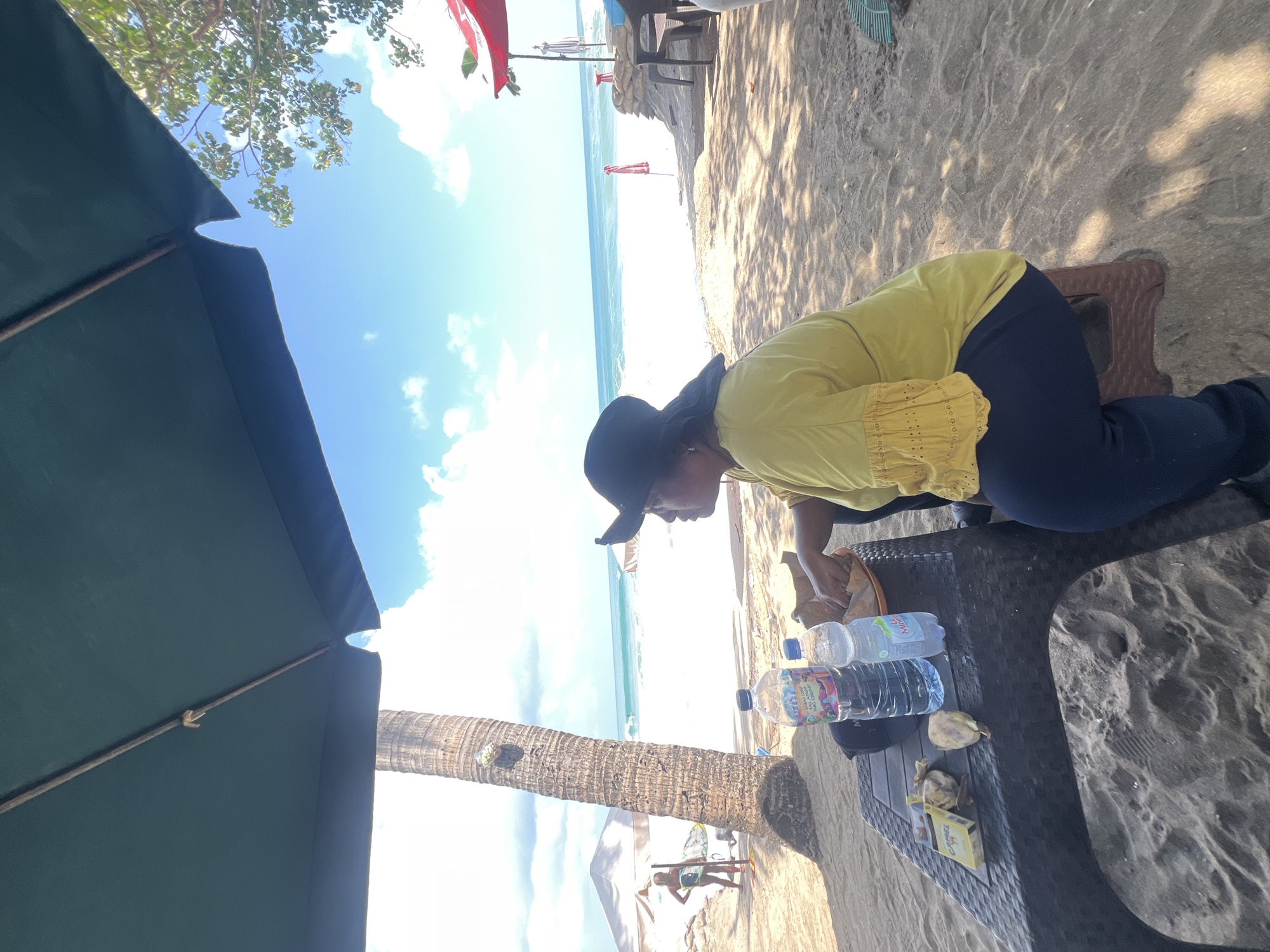 An outdoor scene on a sunny day at Kuta Beach, Indonesia, depicts a woman sitting at a table under a dark green umbrella. The woman, wearing a yellow shirt and a black hat, is facing right, looking out towards the ocean. On the table are two plastic water bottles, a pack of cigarettes, and some food items. A palm tree stands to the left of the table, and the sandy beach stretches towards the turquoise ocean with gentle waves. In the background, other beach umbrellas and structures are visible, along with a person standing near the water's edge. The sky is blue with scattered white clouds.