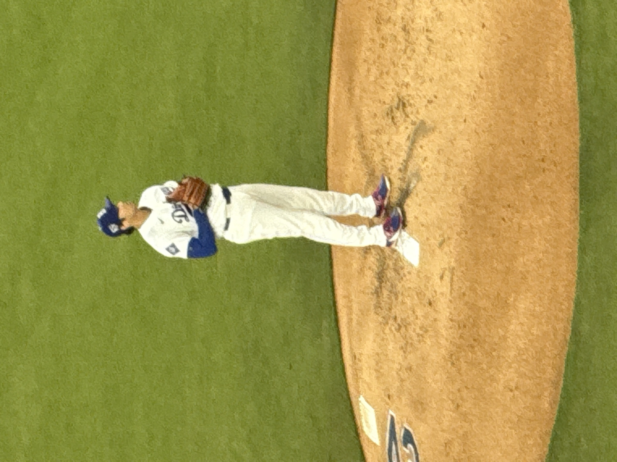 A baseball player in a Dodgers uniform is standing on the pitcher's mound at Dodger Stadium in Los Angeles. The field is green with the pitcher's mound in the center. The player is wearing a glove and baseball cap, ready to throw a pitch.
