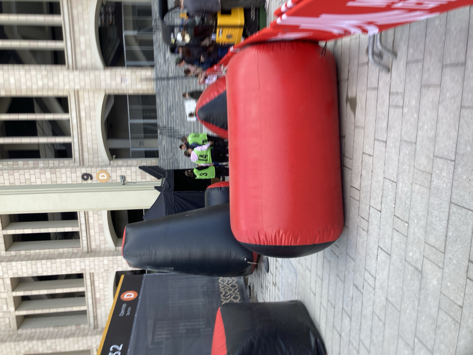 The image shows an outdoor scene with a building on the left and inflatable obstacles in the foreground. The building features multiple windows arranged in a grid pattern, and a sign labeled "D" is visible. People are standing and walking behind the inflatable obstacles. The obstacles are red and black cylindrical shapes. The ground is paved with rectangular stones. The location is Moscow, Russia.