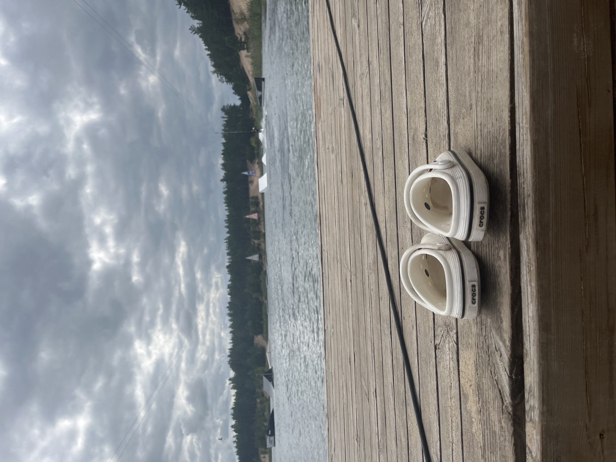 A pair of white Crocs sits on a wooden dock on a cloudy day at the Männiku Wake Park in Männiku, Estonia. The wake park is visible across the lake, with ramps and a wakeboarder in action. The water is choppy, and the sky is filled with dark, heavy clouds. Trees line the distant shore.