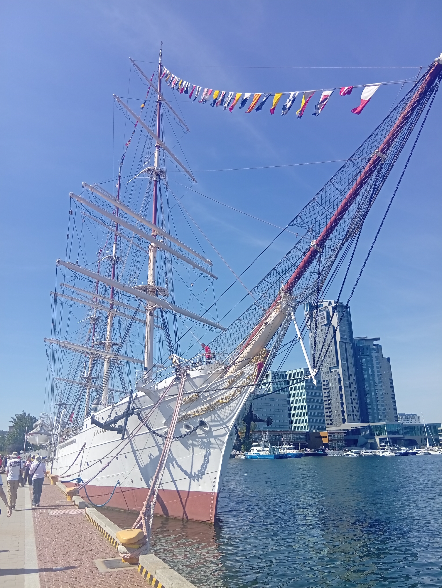 The image shows a large white sailing ship docked in a harbor with modern buildings in the background. The ship has multiple masts and is adorned with a string of colorful maritime flags. The water is calm, and several smaller boats are visible in the distance. People are walking along the quay in the foreground. The architecture of the buildings suggests the location is in Gdynia, Poland, likely at the Gdynia Waterfront or a similar port area known for its historic ships and modern development. The sky is clear and blue, indicating a sunny day.