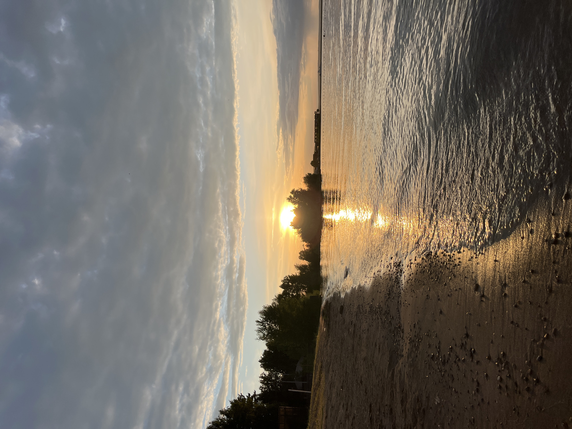 This is a vertical, wide-angle shot of a beach scene at sunset. The sun, a bright orb of yellow and white light, is low on the horizon, casting a warm, golden glow across the water and the sky. The sun's reflection shimmers on the rippling surface of the water, creating a path of light that stretches towards the viewer.
The foreground is dominated by a sandy beach, with small pebbles scattered across its surface. The water's edge laps gently at the shore, with small waves creating a foamy line where they meet the sand.
In the background, a line of trees forms a dark silhouette against the bright sky. Beyond the trees, a faint horizon line suggests a distant land or structures. The sky is a mix of dramatic clouds, with patches of bright sunlight breaking through the grayer, more overcast areas. The overall mood is tranquil and peaceful, evoking a sense of end-of-day serenity.
The time of day is clearly sunset, indicated by the low angle of the sun and its warm, golden light. The weather appears to be partly cloudy, with a mix of sun and cloud. The location is a beach, likely a lake or a calm sea, given the gentle waves. There is no visible text in the image. The scene is natural and serene.