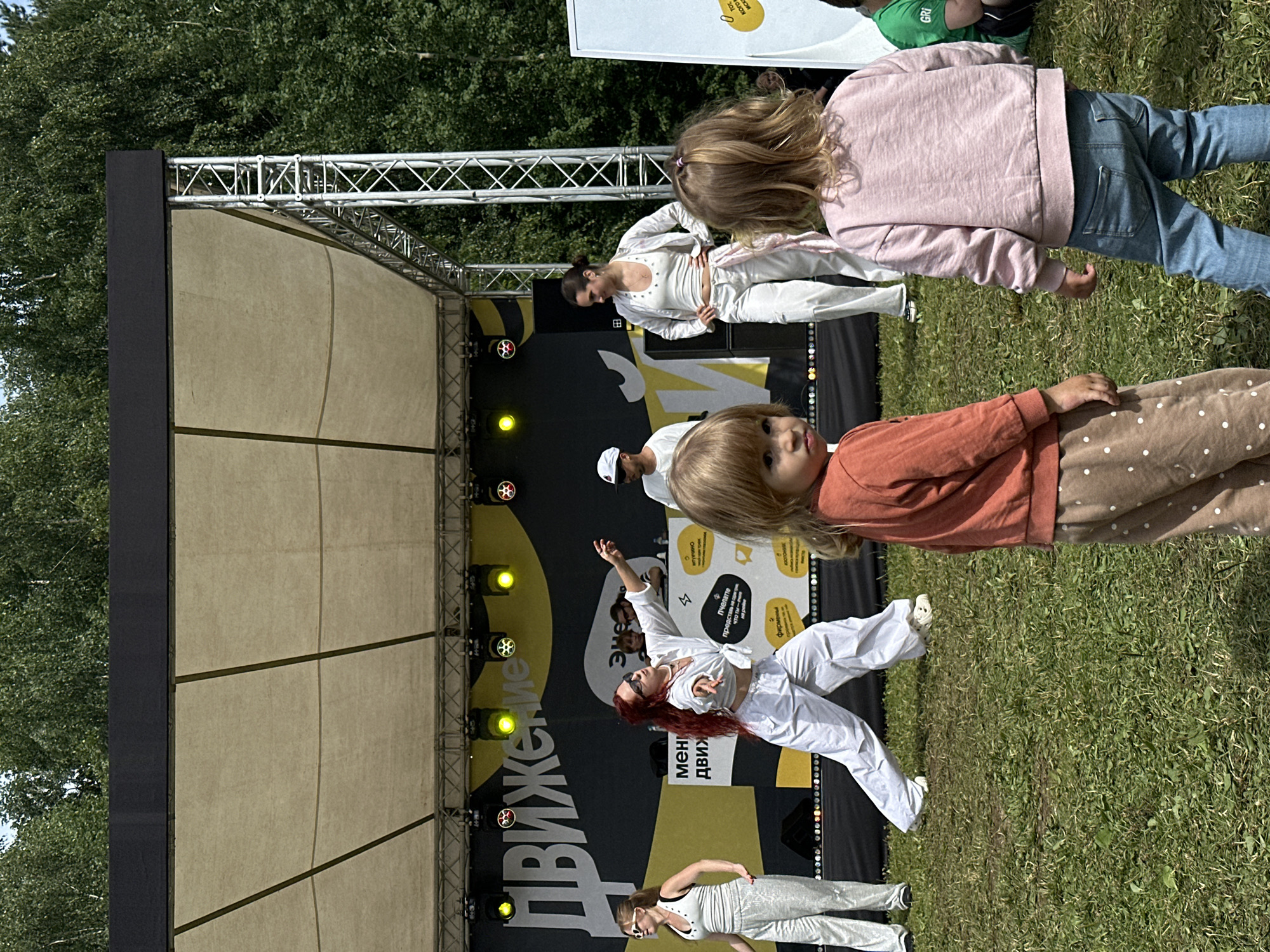 An outdoor stage event is taking place in Moscow, Russia, on a sunny day. A group of people, primarily young women dressed in white athletic-style clothing, are performing or dancing on a stage. The stage has a yellow backdrop with large white lettering that appears to be Russian text, possibly indicating the event's name or theme. Several stage lights are visible, casting a warm glow. In the foreground, two young children, a boy and a girl, are facing the stage, seemingly observing the performance. The ground is covered in green grass, and trees are visible in the background, suggesting a park or outdoor venue.