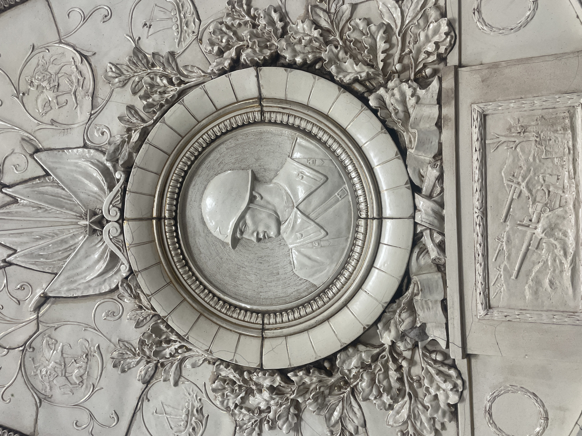 This close-up shows a section of decorative bas-relief wall art. Centered is a circular medallion featuring a left-facing profile of a man in military uniform and helmet. The medallion has a heavily ornamented border with an outer band of square segments. Surrounding the medallion are sculpted oak leaves and acorns. To the left, there's another circular relief with what appears to be equestrian figures. To the right of the central medallion, a rectangular panel shows military imagery, including what looks like crossed rifles or tools. The entire piece is rendered in white, possibly plaster or a similar material. This decorative panel is located in the Moscow, Russia Metro system on the Tagansko-Krasnopresnenskaya line, Slavyansky Bulvar station.