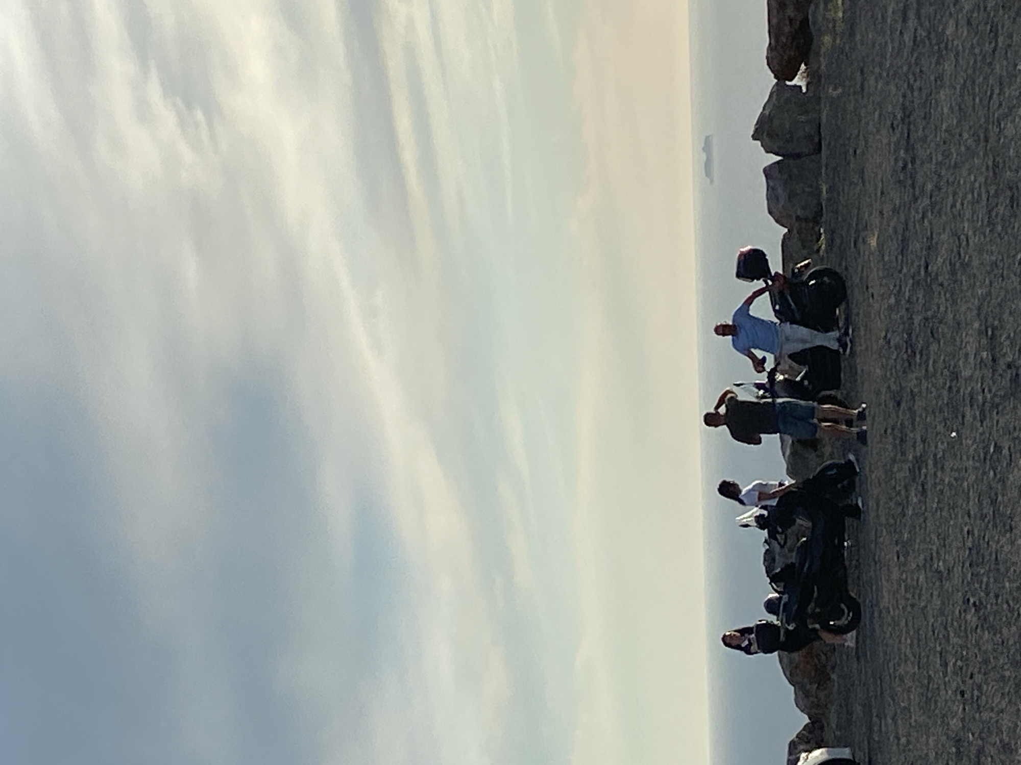 In the city of Kovačko Polje, Montenegro, on a rocky terrain overlooking a vast body of water, a group of four individuals are gathered around two motorcycles. The sky above is a soft blend of blues and whites, with faint clouds drifting across the horizon. The sea stretches out, meeting the sky in a hazy line, and a distant ship is visible on the water.
On the left, a woman with dark hair, wearing a dark top and shorts, stands near a black motorcycle. To her right, a second woman, with dark hair and wearing a white t-shirt and dark pants, leans against the same motorcycle. A man with dark hair, in a dark t-shirt and shorts, stands near the front of the motorcycle, with his hand near his head as if adjusting something or shielding his eyes.
Further to the right, a man in a light blue t-shirt and light-colored pants stands with one hand on the handlebars of a second black motorcycle. This motorcycle has a red storage box on the back. The man appears to be interacting with his companion or the motorcycle. The rocky landscape behind the people and motorcycles is uneven, with large, dark boulders scattered throughout. The ground in the foreground is a mix of dark gravel and small stones.