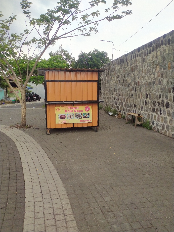 An outdoor scene features an orange-brown metal food cart with a black frame and small wheels, positioned on a paved surface. To the left of the cart stands a tree with green leaves and a slender trunk. To the right, a tall, rough stone wall extends into the distance, made of various dark-colored stones. A small wooden bench sits at the base of the wall on the right side.
The ground is paved with interlocking concrete blocks. In the foreground, a curved section of lighter grey and white blocks creates a distinct path against the darker grey pavement. Beyond the tree, glimpses of parked motorbikes and a building are visible. The sky is overcast, indicating daytime.
The front of the food cart displays a yellow banner with the prominent text "Pangsit Kaki Naga" (Dragon Foot Dumplings/Wontons) in red and white letters. Below this, four circular images showcase different food items offered. Visible details on the banner include a price of "11.000" (likely Indonesian Rupiah) and a "50%" discount icon, along with the text "BOX BOX". No people or active events are present, suggesting a quiet moment in this public space in Karanganyar, Indonesia.