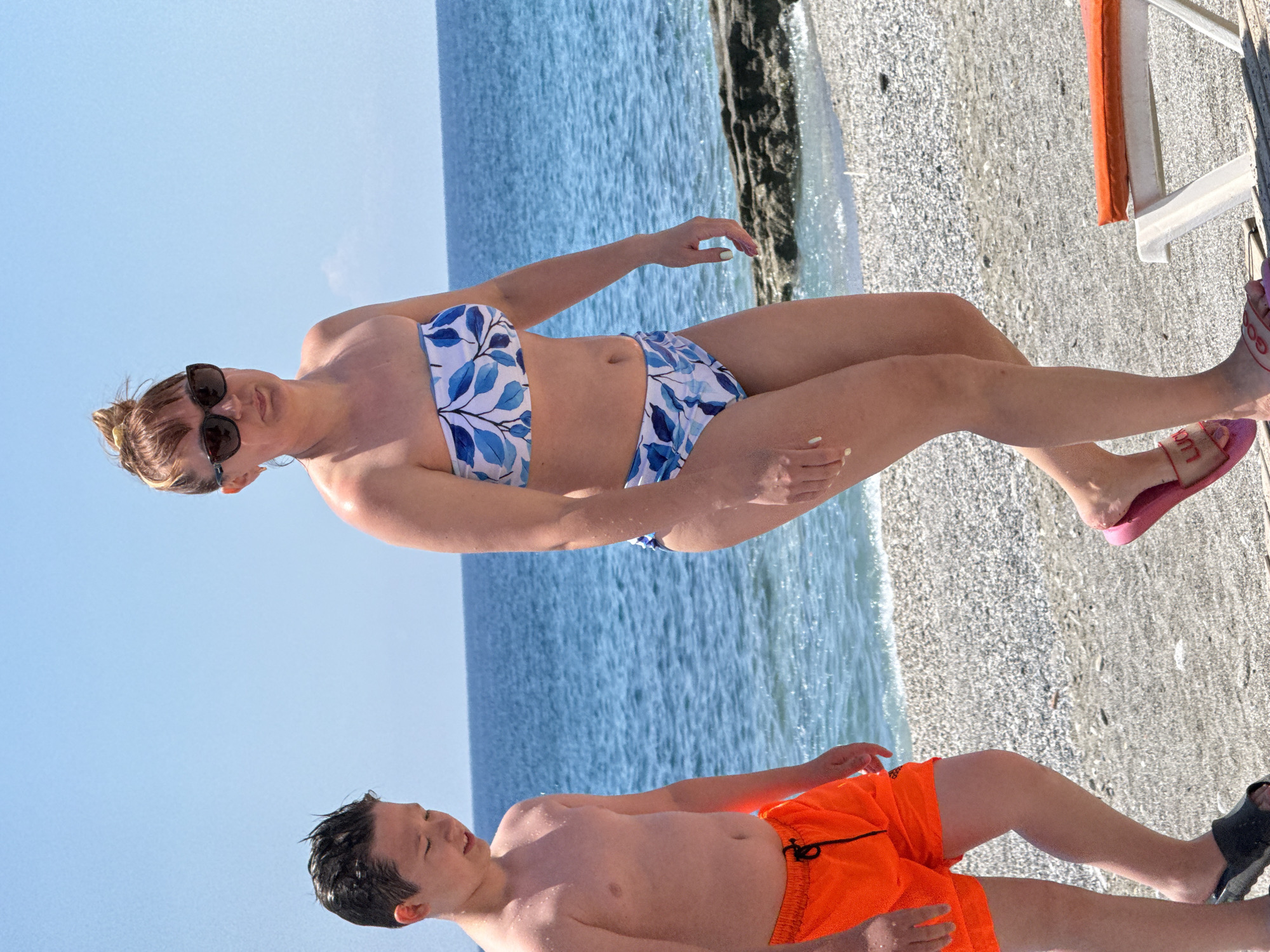 The image, rotated 90 degrees counter-clockwise, depicts two individuals relaxing on a pebble beach beside the ocean under a bright, sunny sky.
In the foreground, a woman is lying on her side, facing the viewer with a relaxed expression. She wears dark sunglasses and a blue and white bikini with a leafy pattern. Her light brown hair is pulled back, and her fingers and toes have light-colored nail polish. On her feet, she wears pink flip-flops, one of which clearly displays the word "LUCKY."
Slightly further back and to the left, a young boy or child is lying on his back, looking upwards. He is wearing bright orange swim trunks, and his dark hair appears wet. Partially visible on his feet are black sandals.
The background features clear, blue ocean water with gentle waves reaching the pebble shore. The beach itself is covered with small, light-colored stones. A white and orange beach chair or lounger is partially visible on the right side of the image, near the woman's feet. The overall scene conveys a sense of calm and enjoyment on a sunny day at the beach.