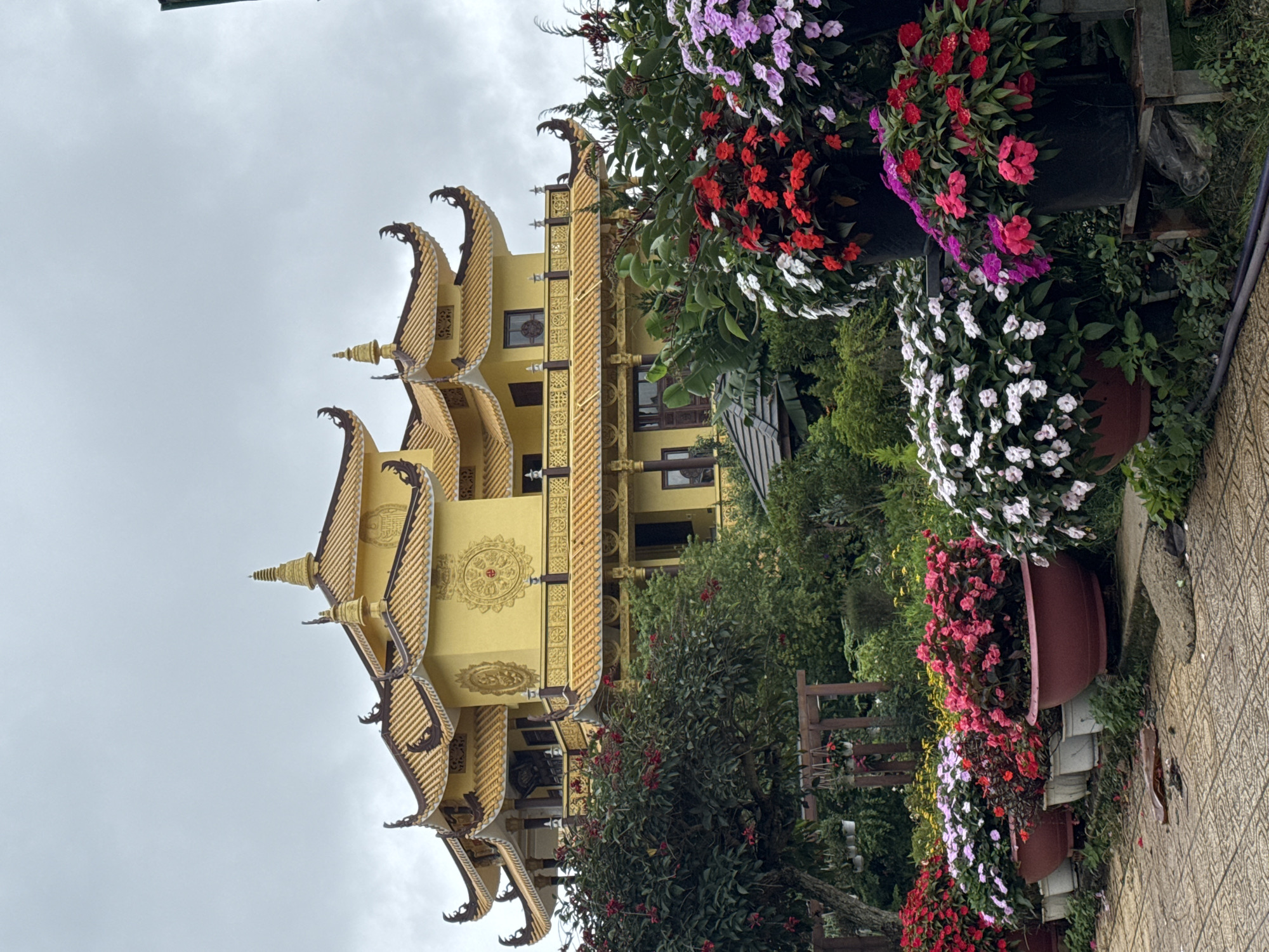 The image shows a tall, yellow building with a multi-tiered, ornate roof in Da Lat, Vietnam. The building is situated amongst greenery and is partially obscured by trees and flower pots filled with red, pink, purple, and white flowers. The sky is overcast. The building has intricate gold detailing on its exterior. A stone-paved path is visible in the foreground.